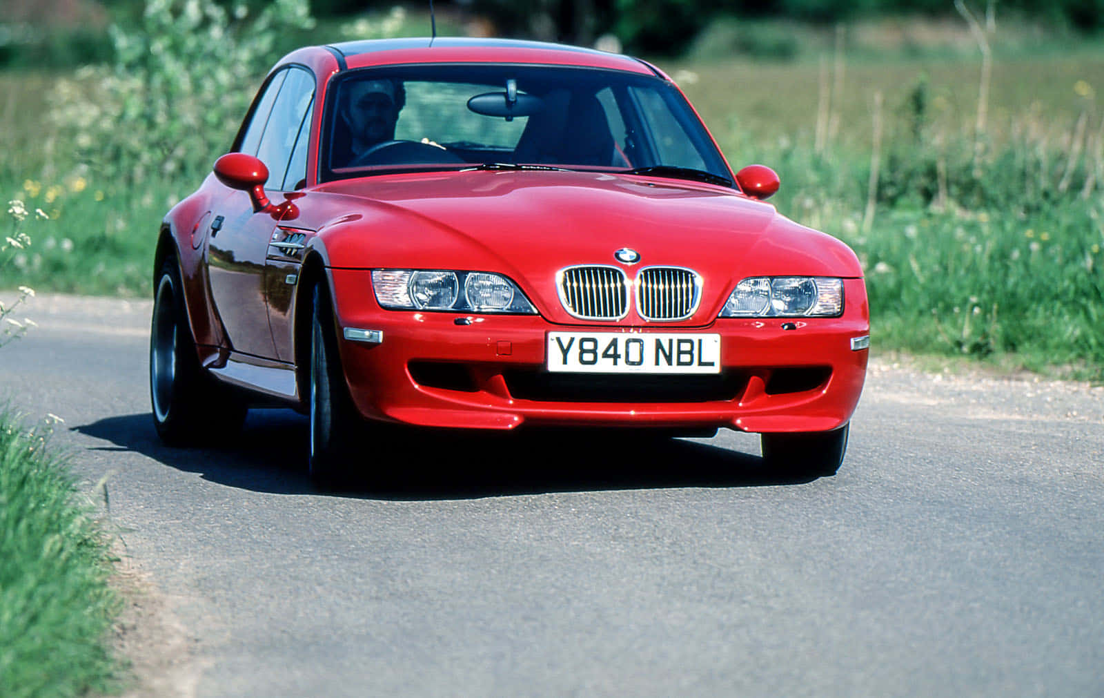 Bmw Z3 桌布 壁紙 — 一輛鮮紅色的BMW Z8跑車在陽光明媚的鄉村道路上行駛，兩旁是綠草和野花。