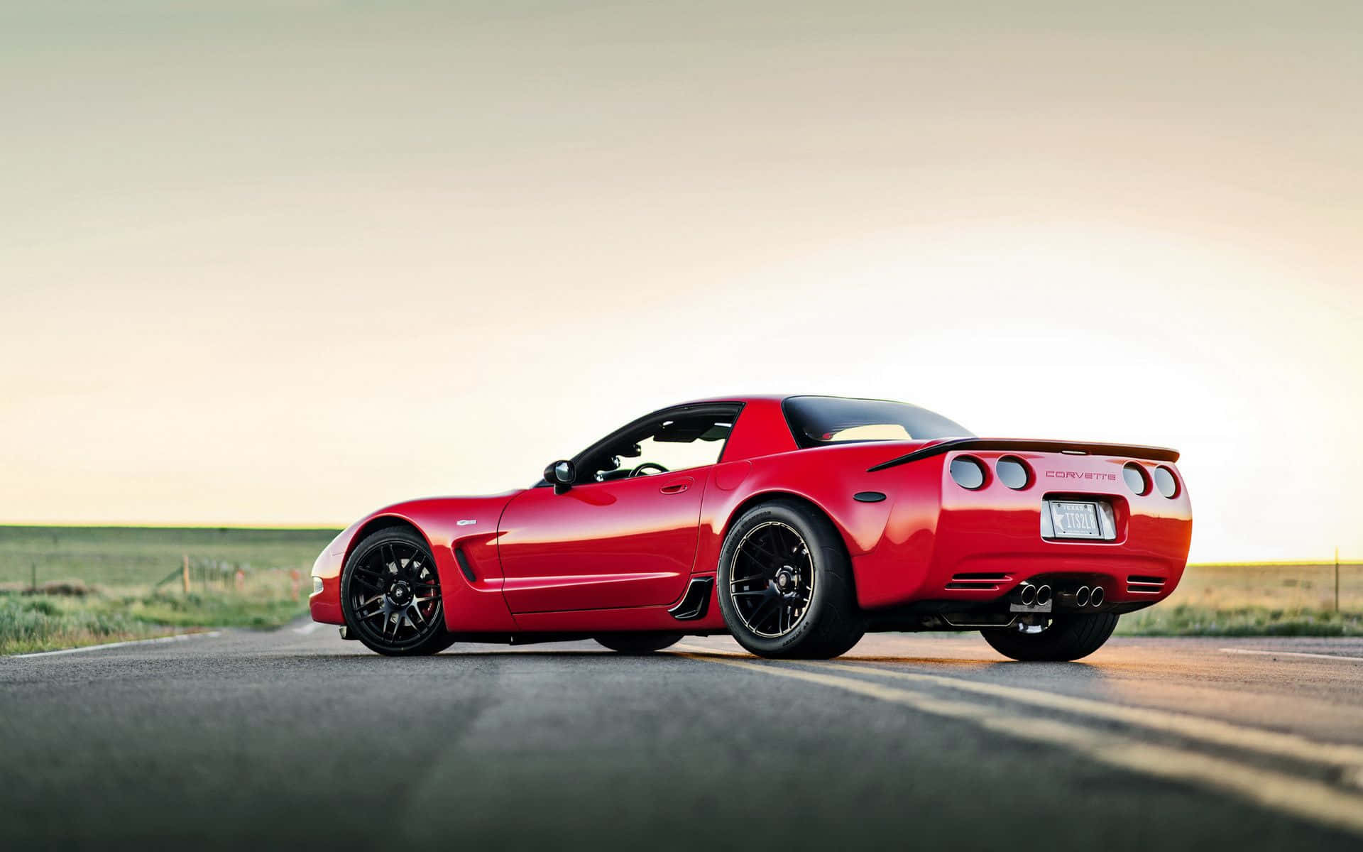 corvette 桌布 壁紙 — 一輛鮮紅色的Corvette跑車在黃金時段沿著鄉村瀝青路滑行，柔和陽光突顯其流線曲線和黑色車輪。