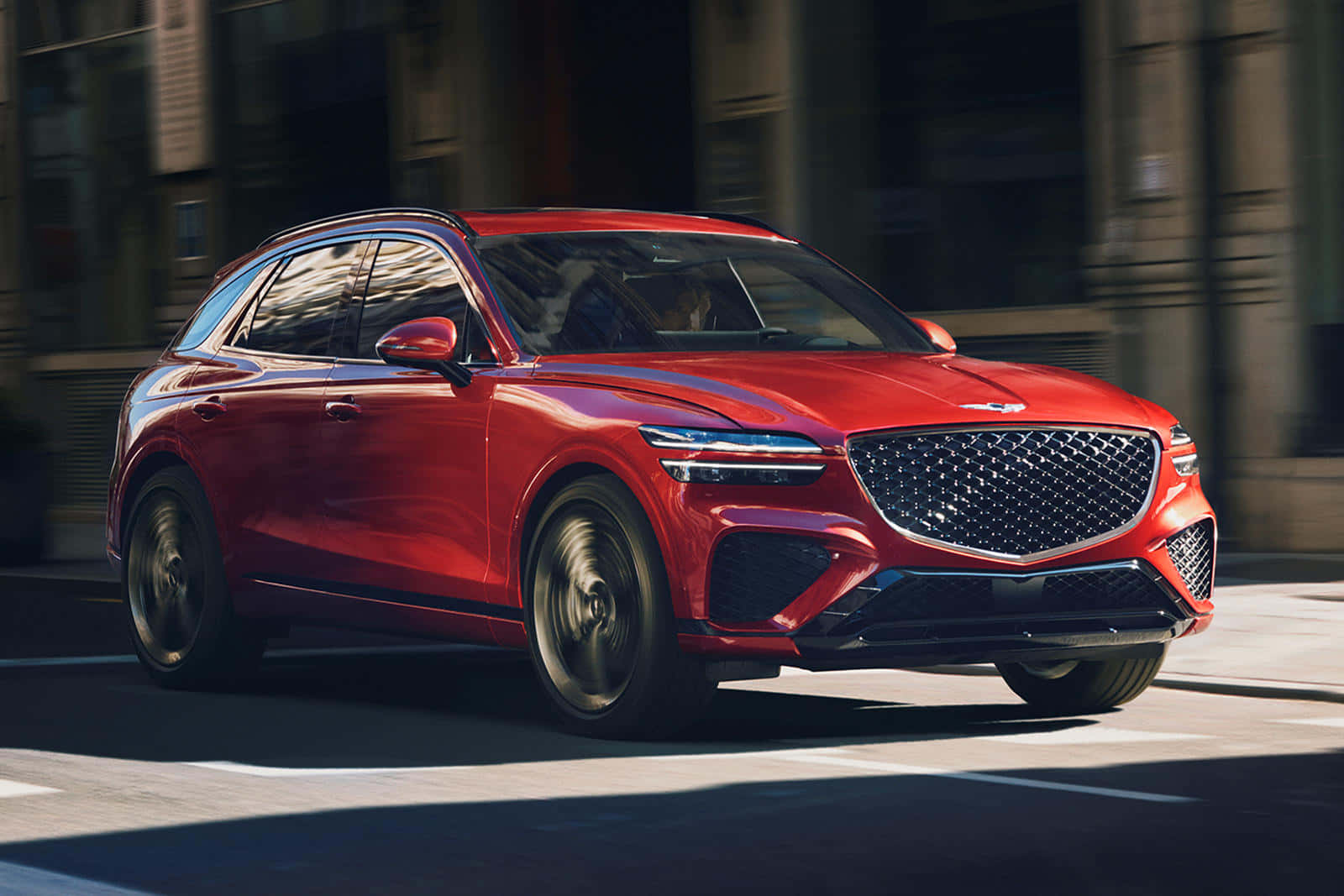 Genesis Gv70 桌布 壁紙 — 一輛鮮紅色的Genesis GV70 SUV在陽光明媚的城市街道上疾馳，車輪和背景建築物呈現動態模糊效果。