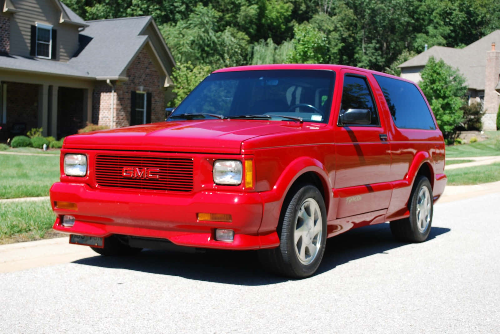 Gmc Typhoon 桌布 壁紙 — 一輛鮮紅色的GMC Typhoon運動型多功能車停放在郊區街道上，背景有房屋和綠樹。
