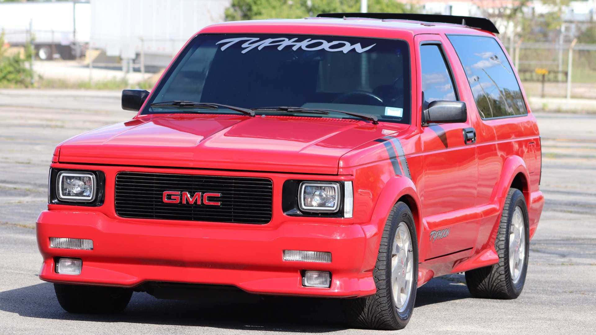 Gmc Typhoon 桌布 壁紙 — 一輛鮮紅色的GMC Typhoon SUV停在瀝青路面上，帶有醒目的黑色賽車條紋和擋風玻璃上的「TYPHOON」貼紙。