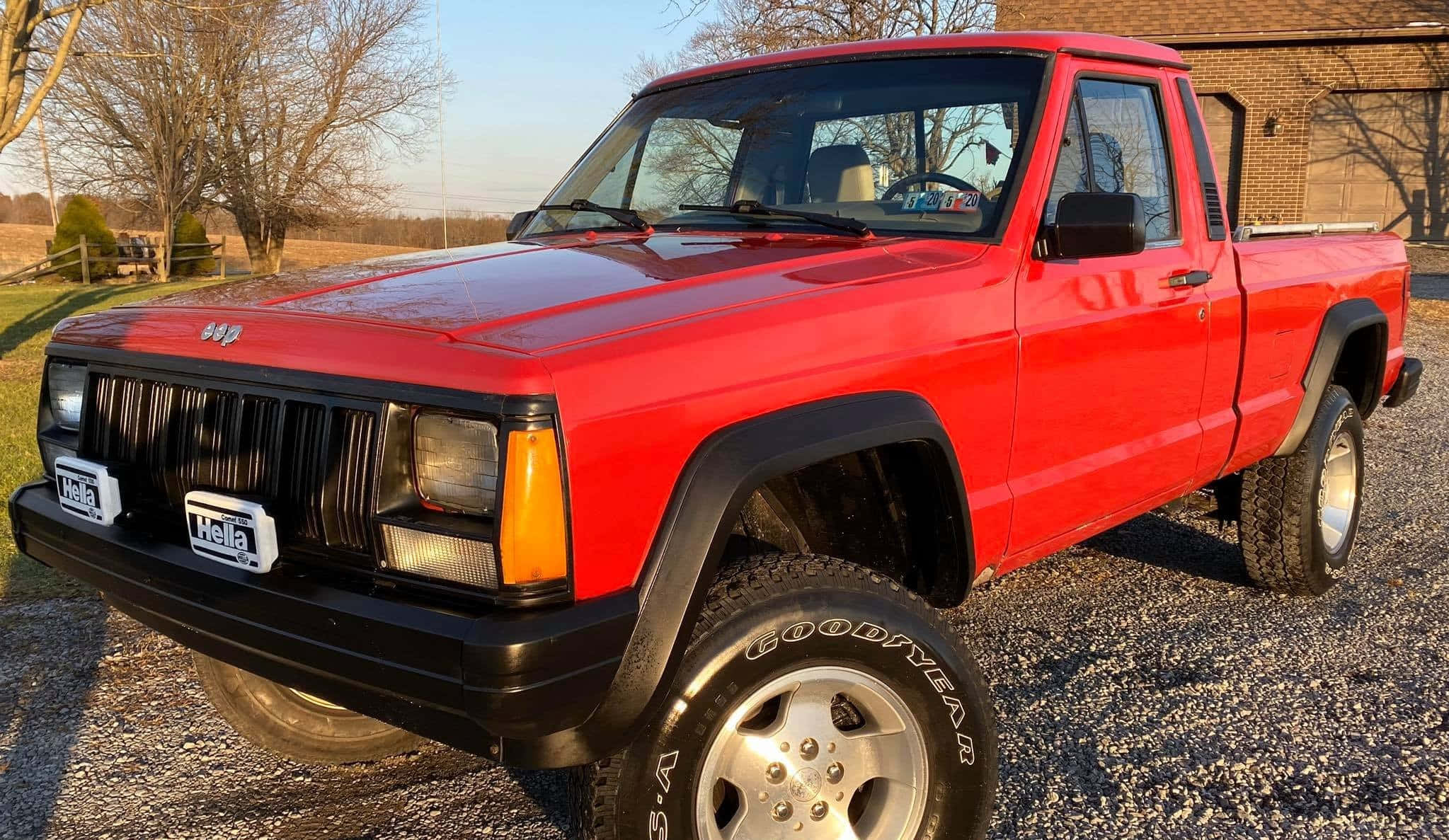Jeep Comanche 桌布 壁紙 — 一輛鮮紅色的Jeep Cherokee皮卡車停在碎石上，背景有光禿的樹木和磚砌建築，天空晴朗。