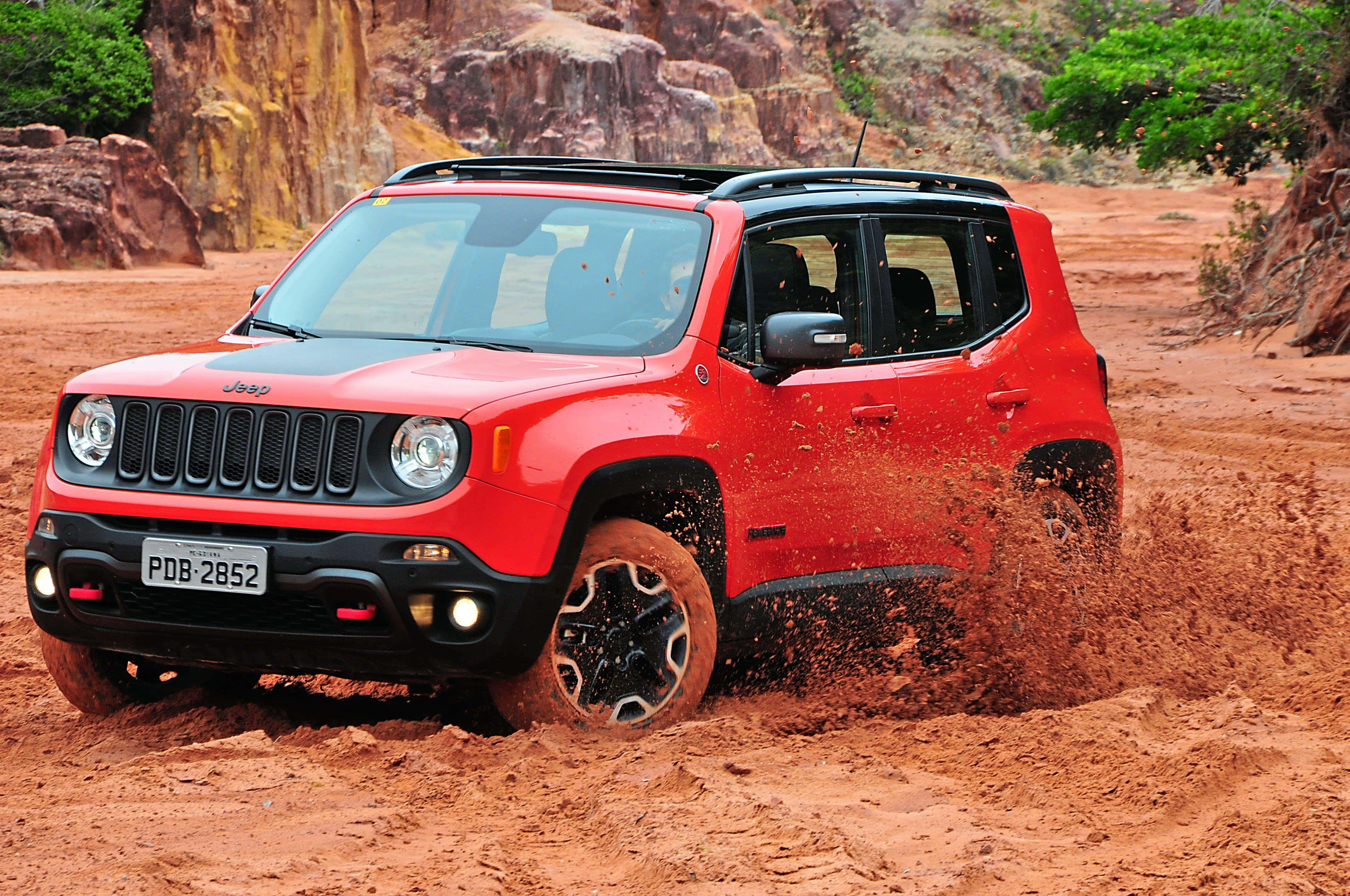 Jeep Renegade 桌布 壁紙 — 一輛鮮紅色的Jeep Renegade在崎嶇的沙漠峽谷中行駛，揚起紅色塵土，周圍有岩石峭壁和綠樹。
