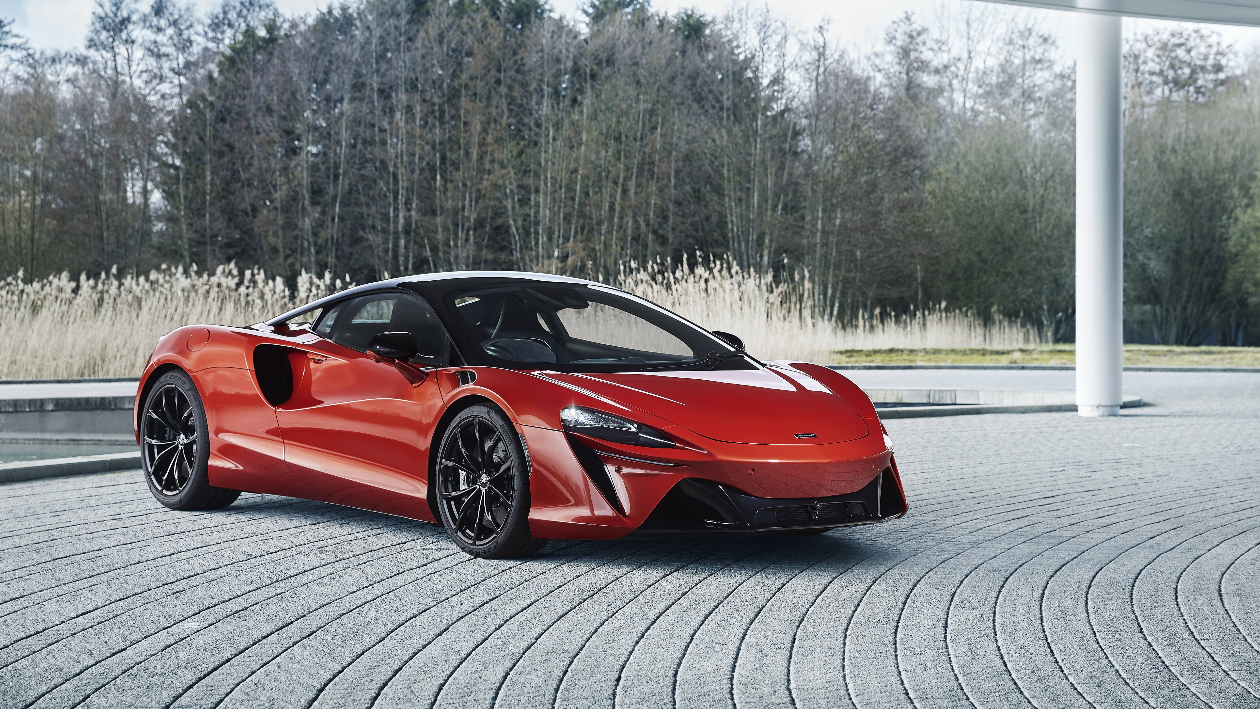Sports Car 桌布 壁紙 — 一輛鮮紅色的McLaren跑車在日光下閃耀，背景是彎曲的石頭庭院，周圍有蘆葦和樹木。