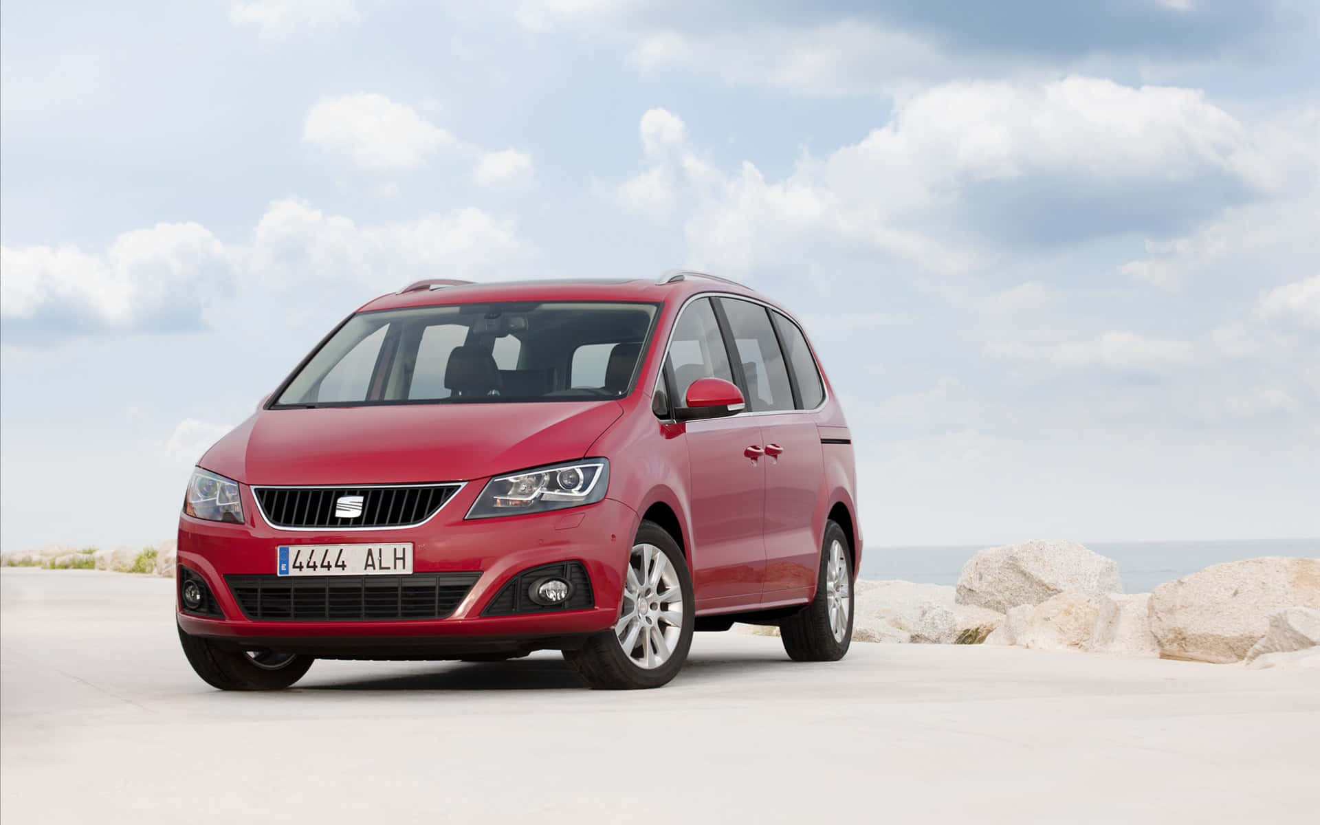 seat 桌布 壁紙 — 一輛鮮紅色的SEAT Alhambra小型貨車停泊在陽光明媚的海岸道路上，周圍散落著岩石，天空多雲。