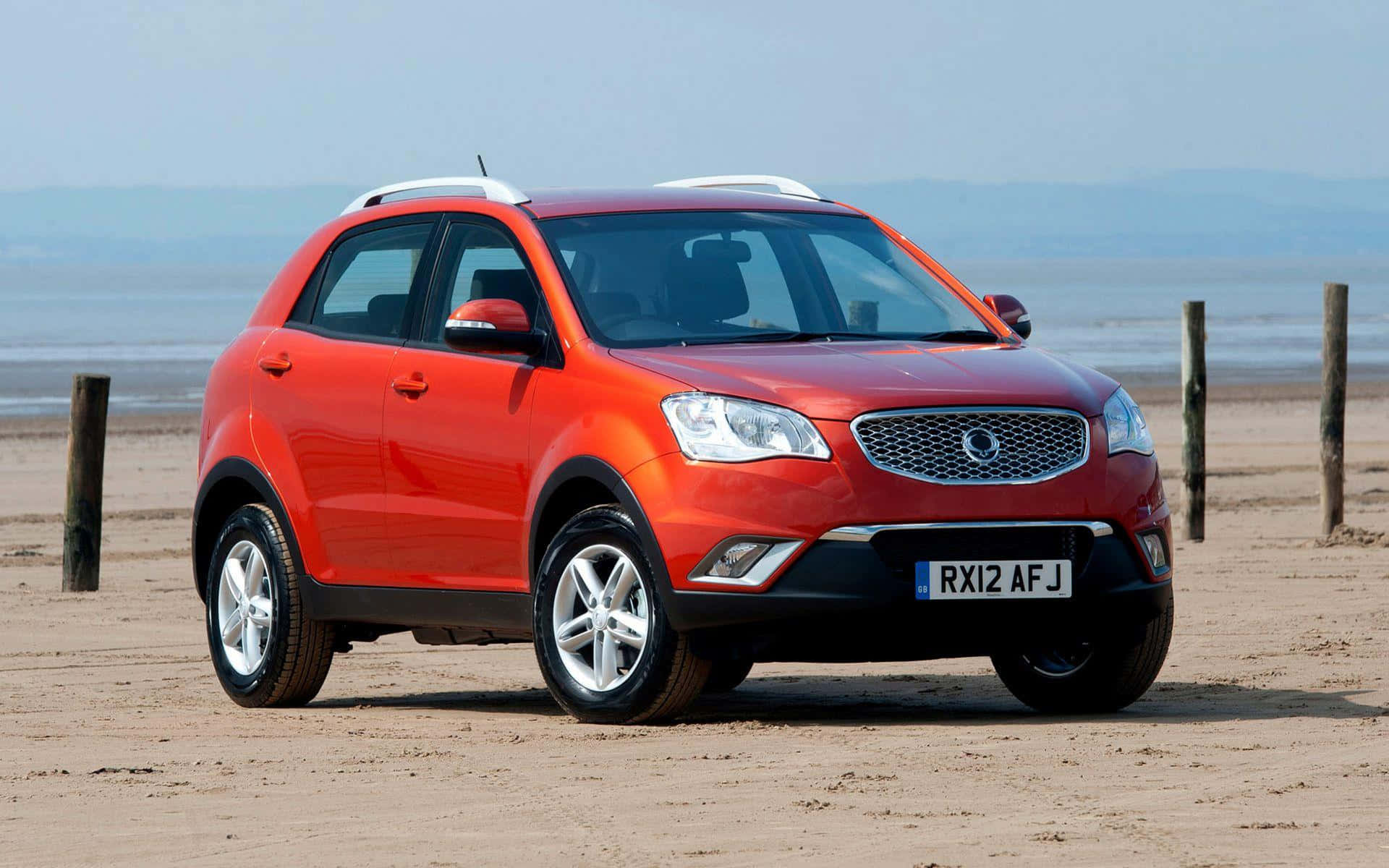 Ssangyong 桌布 壁紙 — 一輛鮮紅色的SUV停泊在沙灘上，背景有木樁和模糊的海岸線。