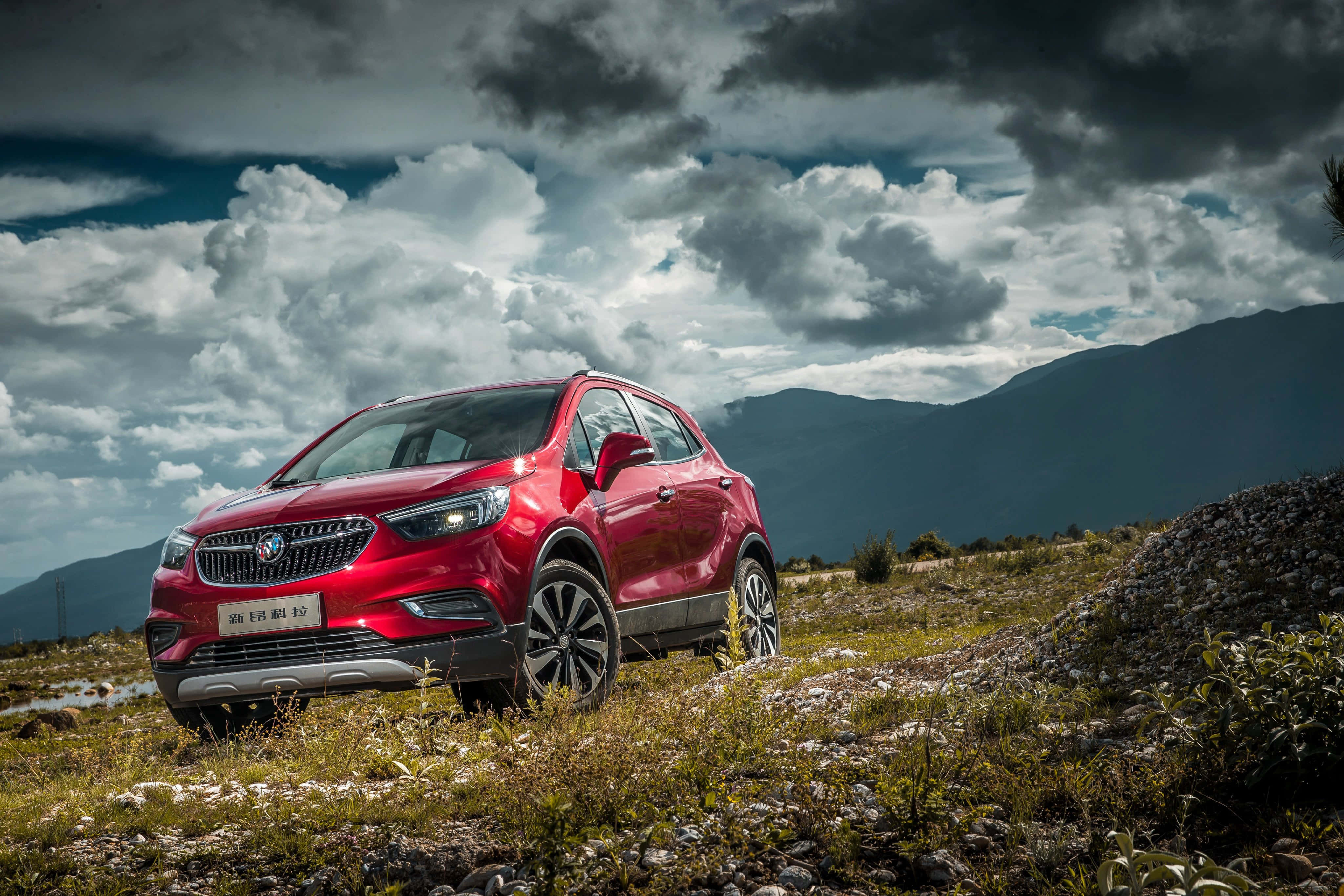 Opel Mokka 桌布 壁紙 — 一輛鮮紅色的SUV在戲劇性的多雲天空下，矗立在崎嶇的草坡上，遠方有山脈。