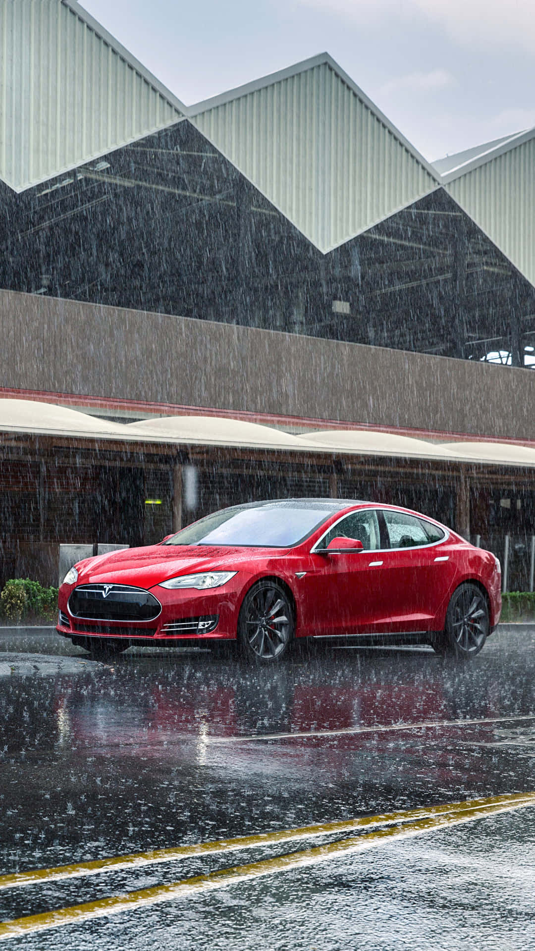 tesla 桌布 壁紙 — 一輛鮮紅色的Tesla房車在滂沱大雨中閃閃發光，背景是現代工業建築的濕漉漉的城市街道。