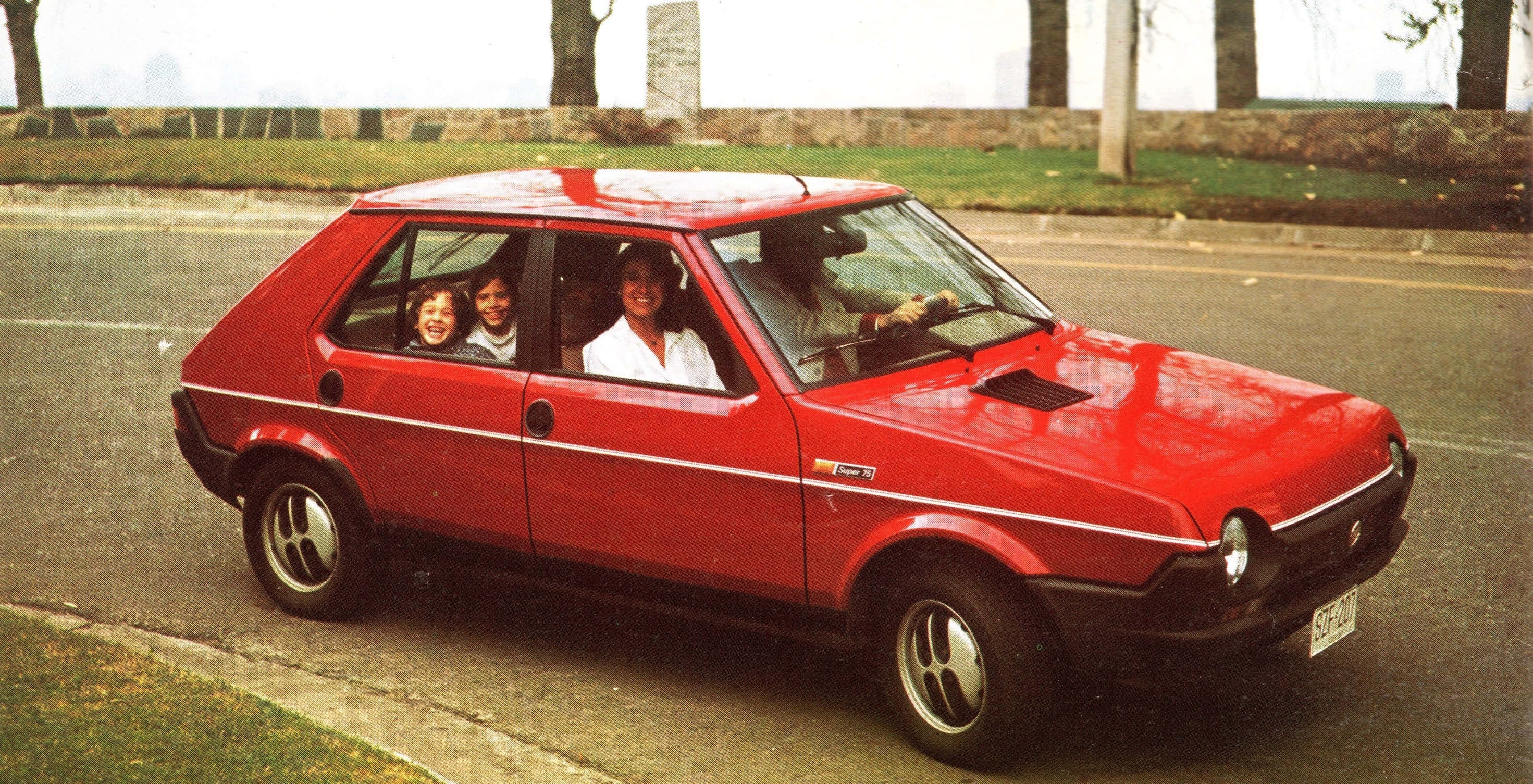 Fiat Ritmo 桌布 壁紙 — 一輛鮮紅色的復古掀背車，車上四位笑容滿面的乘客在晴朗的日子裡行駛在綠樹成蔭的街道上。