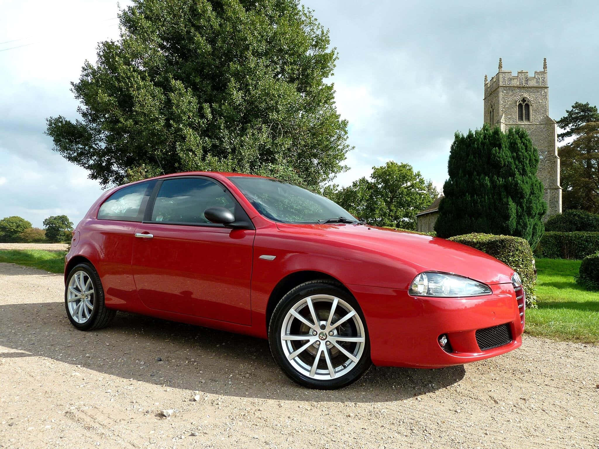Alfa Romeo 147 桌布 壁紙 — 一輛鮮紅色的愛快羅密歐147停泊在碎石車道上，背景是石造教堂塔樓和茂密的綠樹，天空多雲。