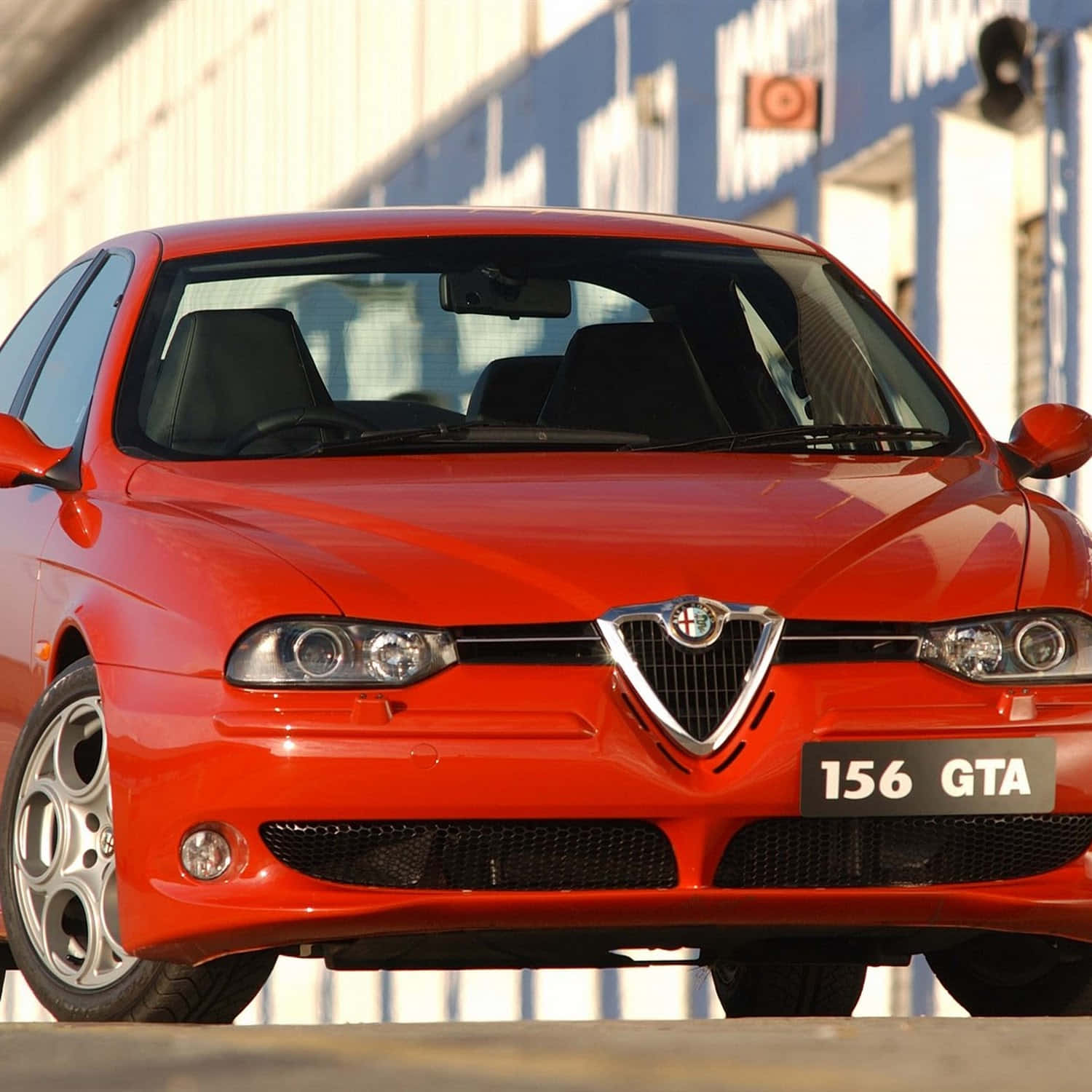 Alfa Romeo 156 桌布 壁紙 — 一輛鮮紅色的愛快羅密歐156 GTA跑車在明亮的陽光下閃閃發光，焦點集中在標誌性的鬼面罩和時尚的頭燈上。