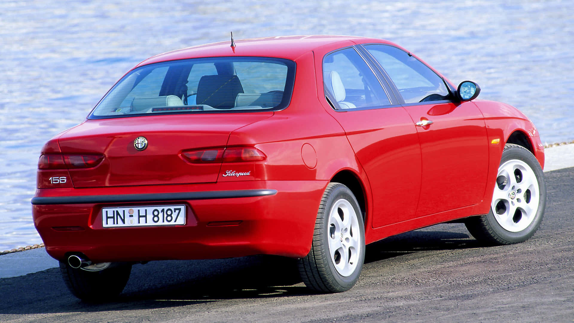 Alfa Romeo 156 桌布 壁紙 — 一輛鮮紅色的愛快羅密歐156轎車停在波光粼粼的水邊，在明亮的日光下。
