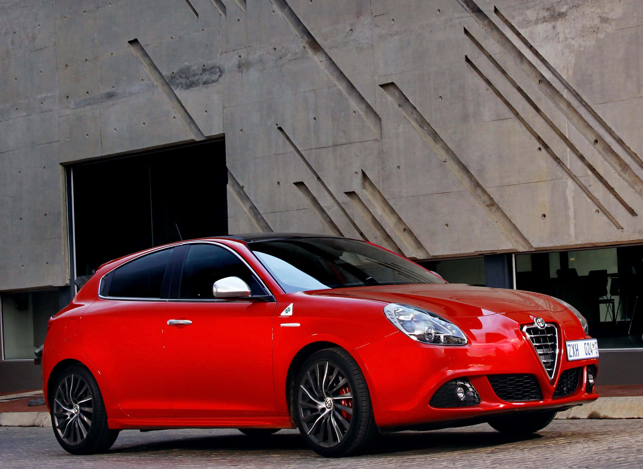 Alfa Romeo Giulietta 桌布 壁紙 — 一輛鮮紅色的愛快羅密歐Giulietta跑車停泊在現代幾何線條的混凝土建築前。