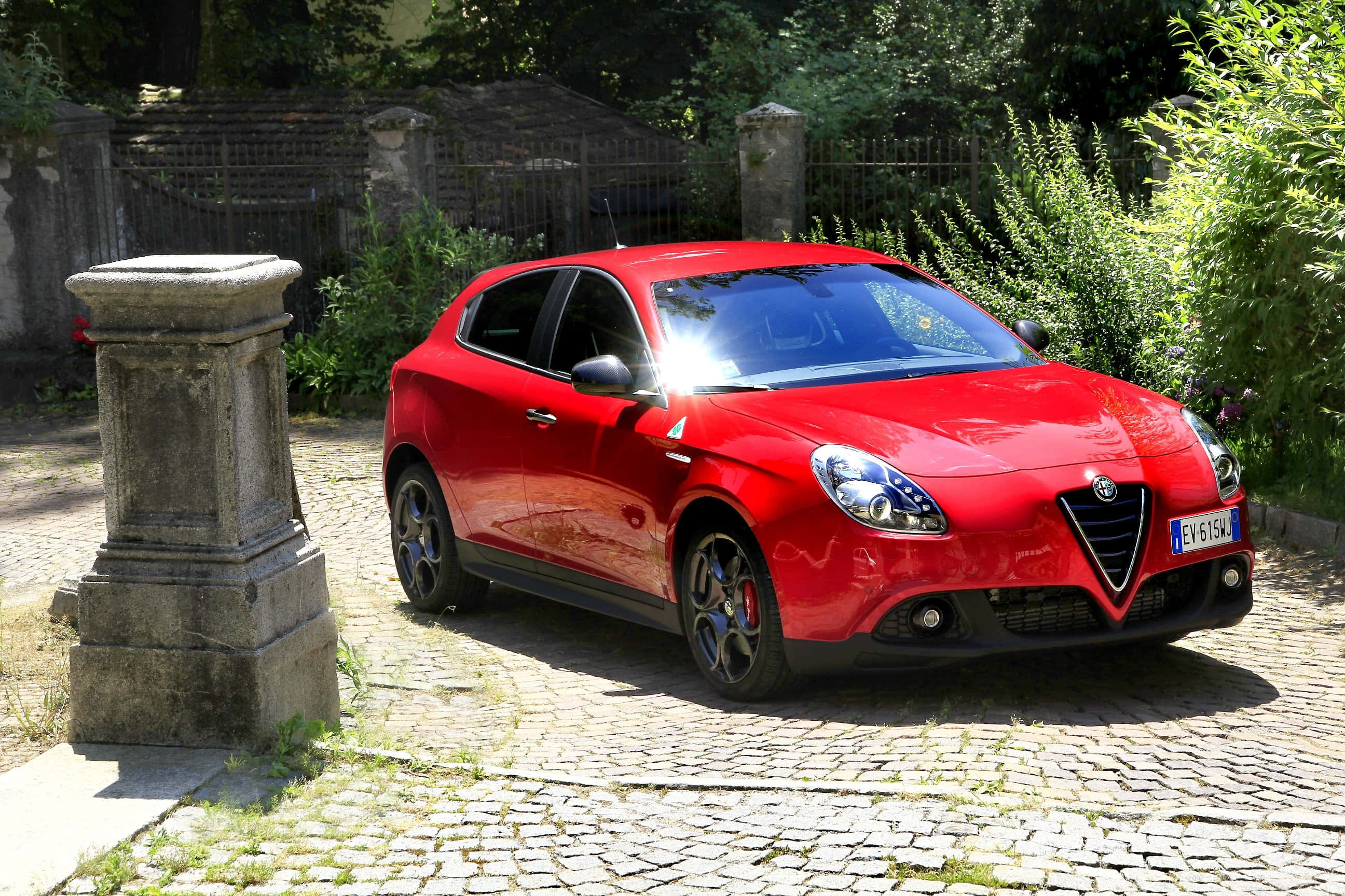 Alfa Romeo Giulietta 桌布 壁紙 — 一輛鮮紅色的愛快羅密歐Giulietta停泊在陽光明媚的鵝卵石車道上，旁邊是一根風化的石柱。
