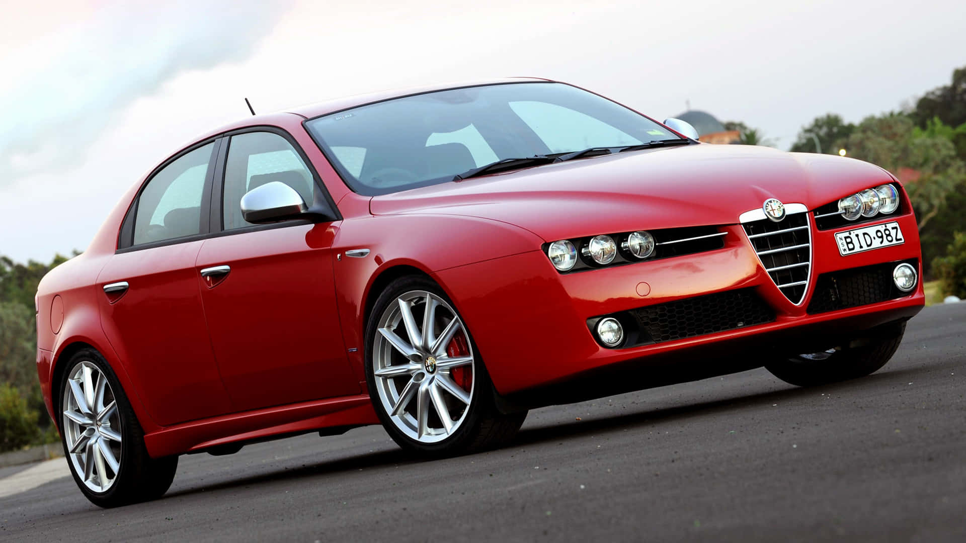 Alfa Romeo 159 桌布 壁紙 — 一輛鮮紅色的愛快羅密歐房車在柔和日光下閃閃發光，停在鋪砌的道路上，背景有綠化。