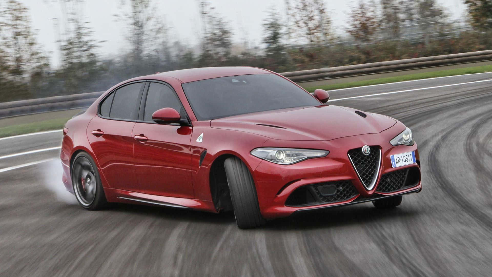 Alfa Romeo Giulia 桌布 壁紙 — 一輛鮮紅色的愛快羅密歐房車在潮濕的賽道上飄移，後輪冒出煙霧，周圍樹木環繞。