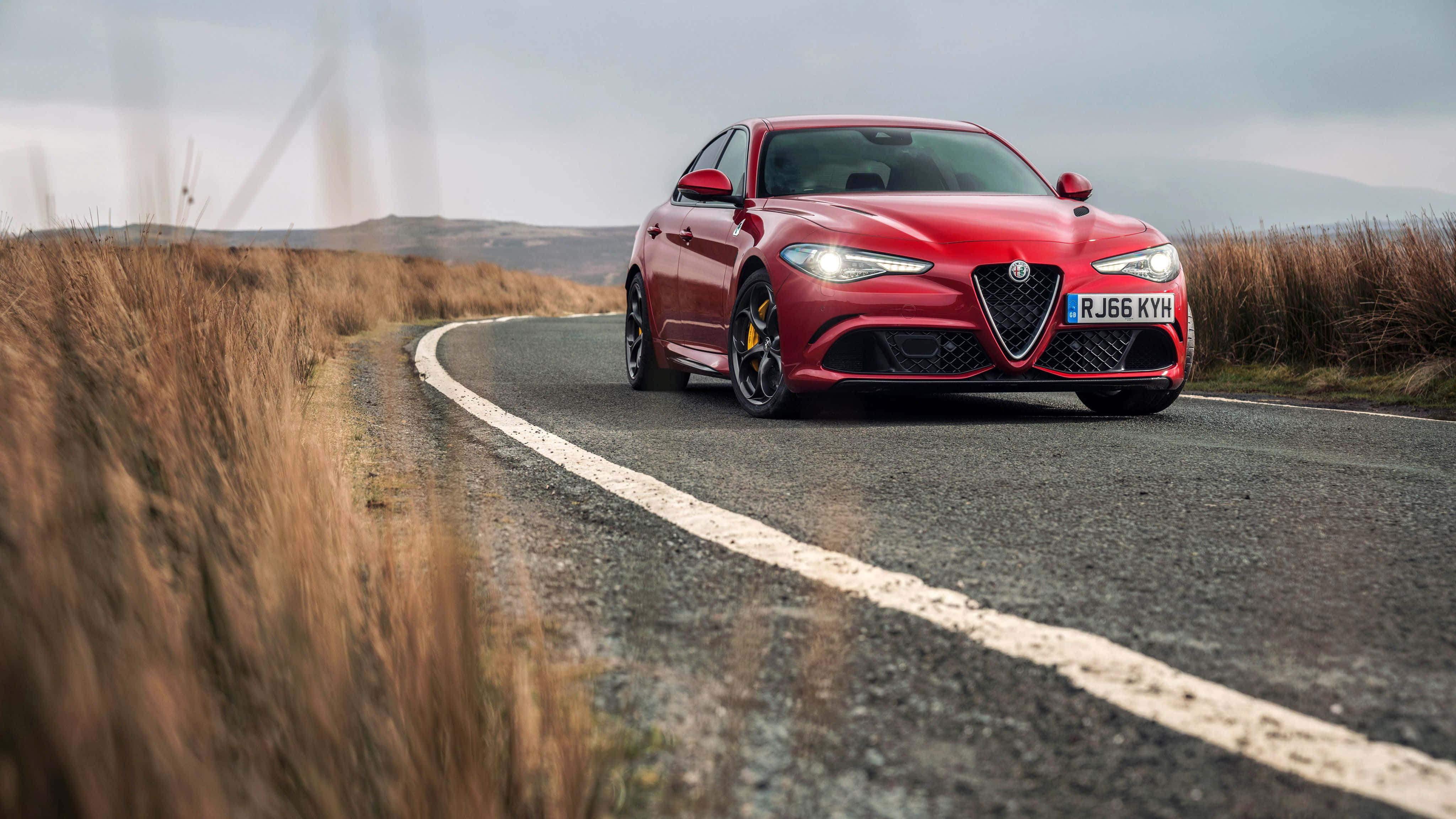 Alfa Romeo Giulia 桌布 壁紙 — 一輛鮮紅色的愛快羅密歐跑車沿著蜿蜒的鄉村道路行駛，兩旁是金黃色的草地，天空多雲。