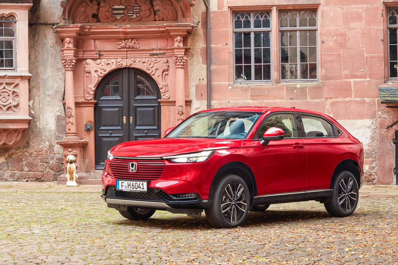 Honda Hr-v 桌布 壁紙 — 一輛鮮紅色的本田SUV停在鵝卵石庭院前，背景是一座帶有華麗雕刻的歷史粉紅色石建築。