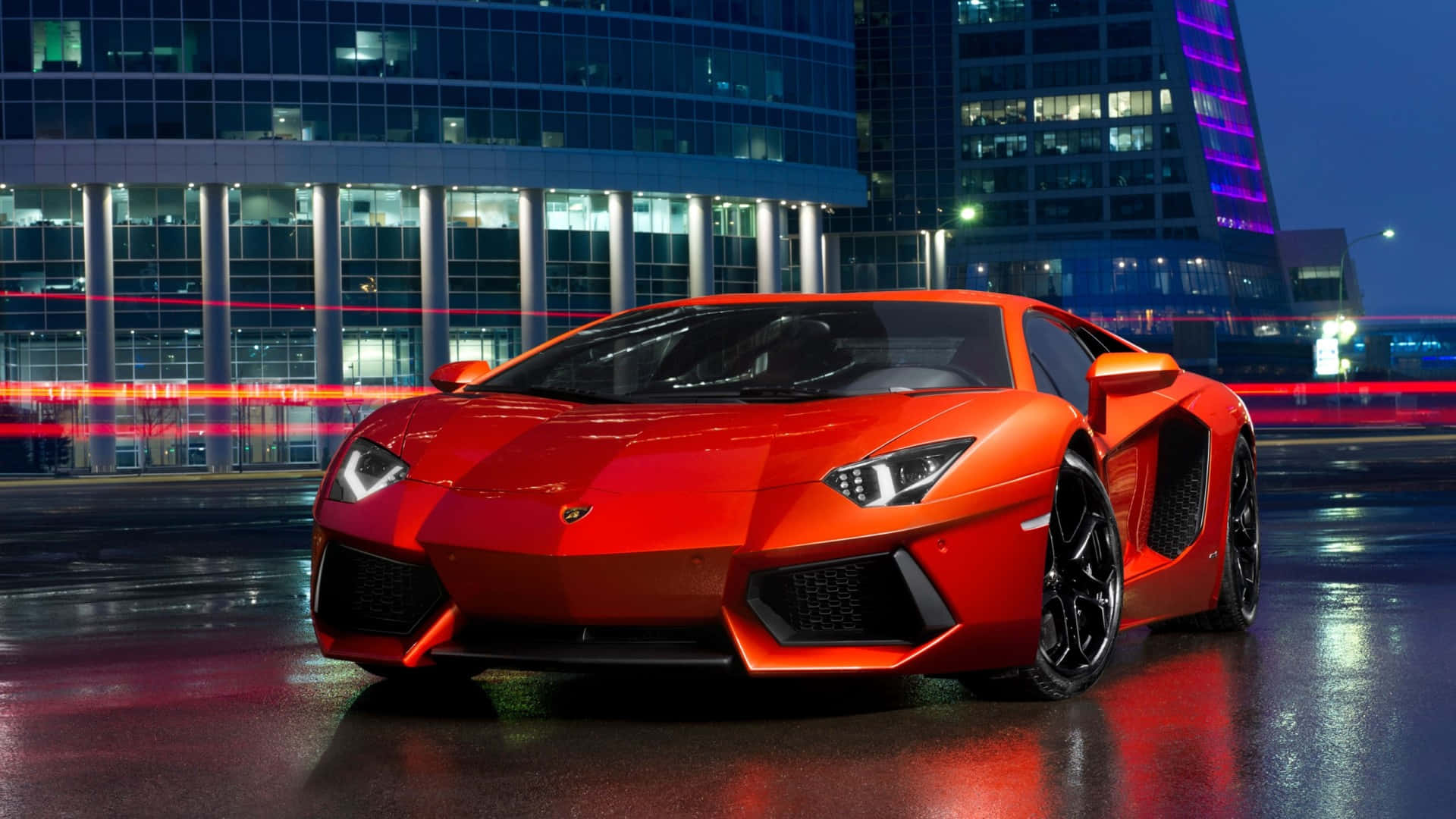 HD Car 桌布 壁紙 — 一輛鮮紅色的林寶堅尼Aventador在都市燈光下，於濕漉漉的夜晚街道上閃耀，背後現代玻璃建築映出模糊的車流痕跡。