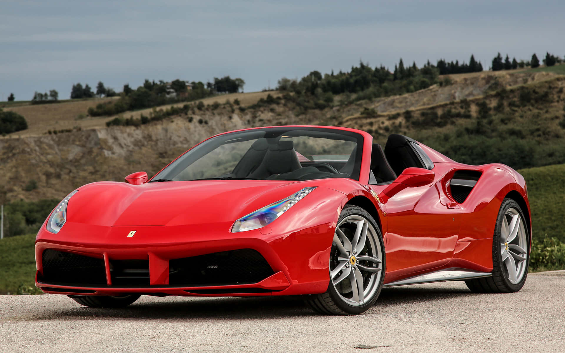Ferrari 488 Spider 桌布 壁紙 — 一輛鮮紅色的法拉利 488 Spider 敞篷車停泊在碎石路上，背景是起伏的丘陵和稀疏的樹木，天空多雲。