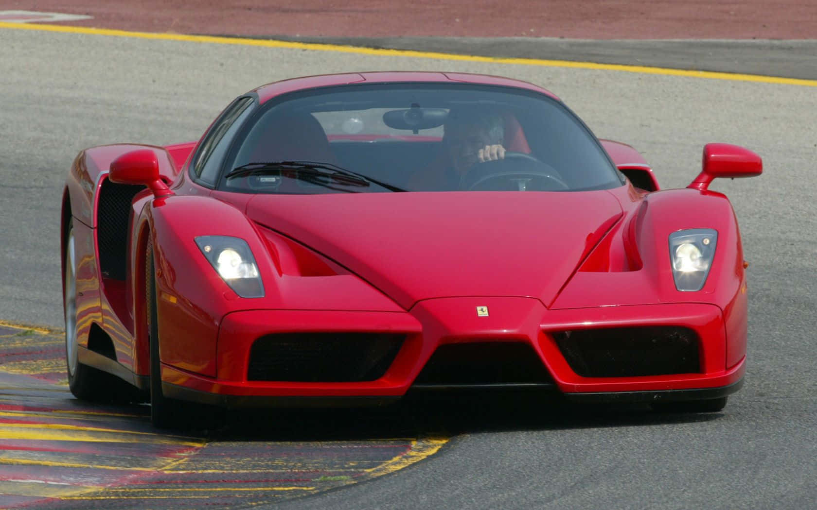 Ferrari Enzo 桌布 壁紙 — 一輛鮮紅色的法拉利 Enzo 在明亮的日光下，沿著帶有黃色路緣石的賽道飛馳。