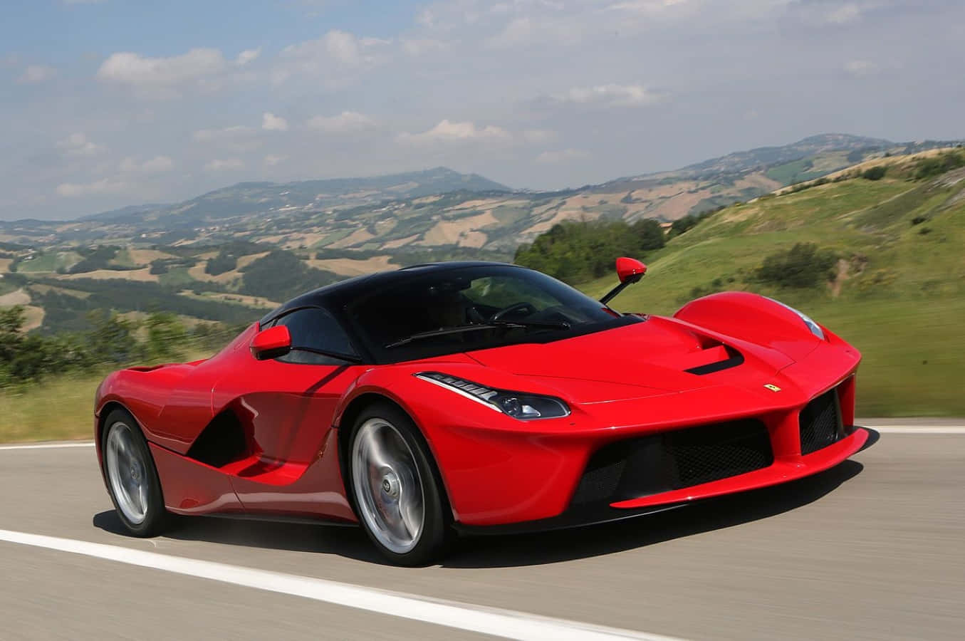 Ferrari Laferrari 桌布 壁紙 — 一輛鮮紅色的法拉利 LaFerrari 在蜿蜒的山路上疾馳，背景是鬱鬱蔥蔥的綠色山丘和部分多雲的天空。