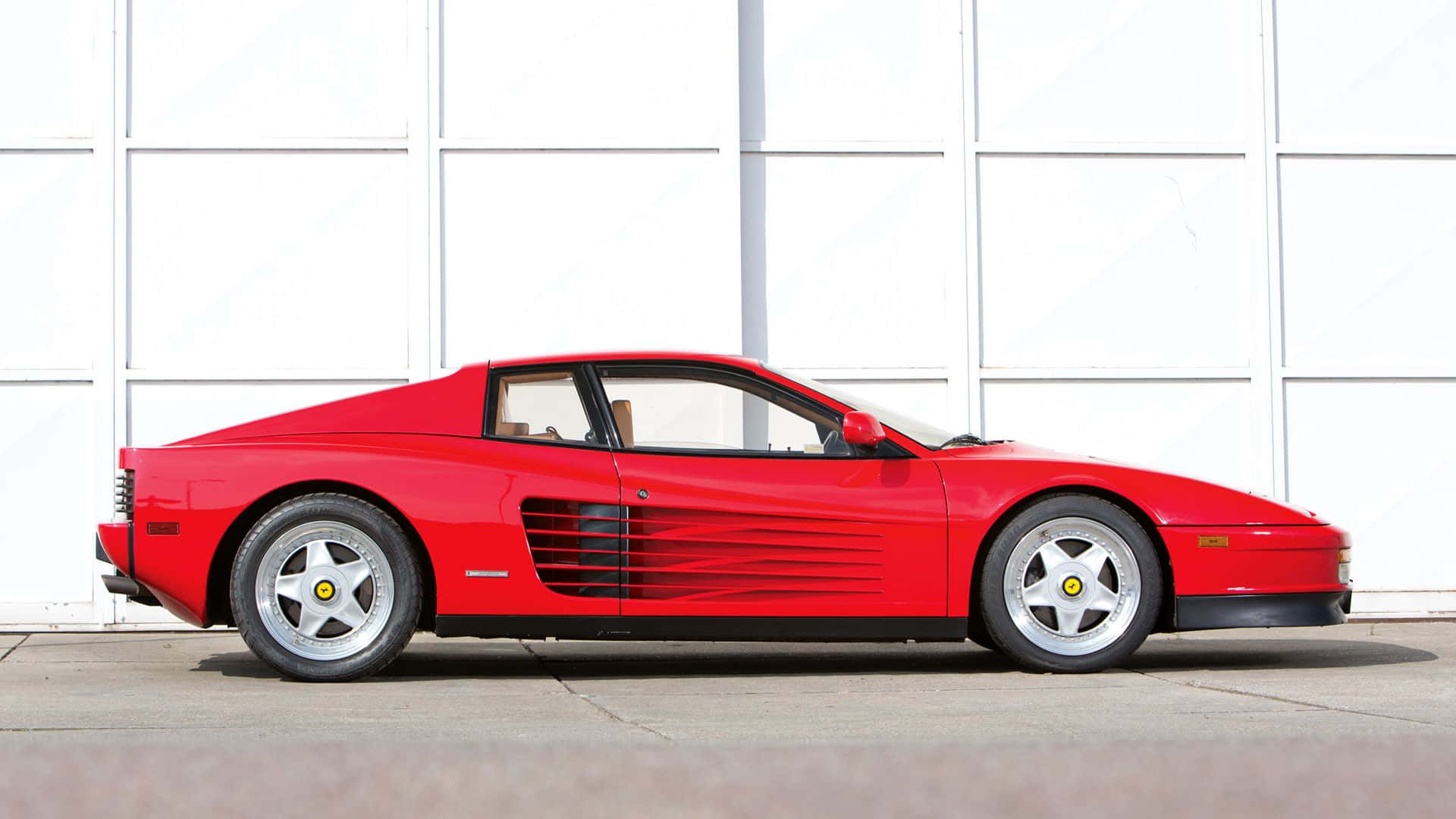 Ferrari Testarossa 桌布 壁紙 — 一輛鮮紅色的法拉利 Testarossa 停放在白色車庫門前，展示其標誌性的側面輪廓和流線型設計。