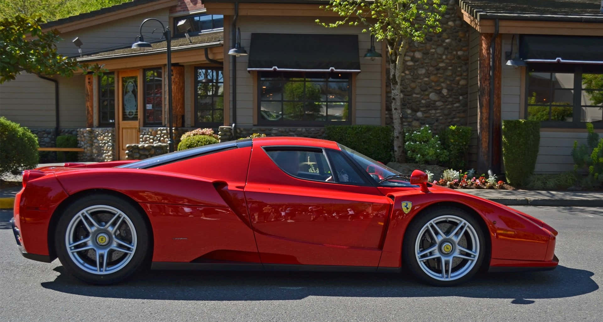 Ferrari Enzo 桌布 壁紙 — 一輛鮮紅色的法拉利Enzo停泊在陽光明媚的街道上，旁邊是一座帶有綠植的質樸石木建築。