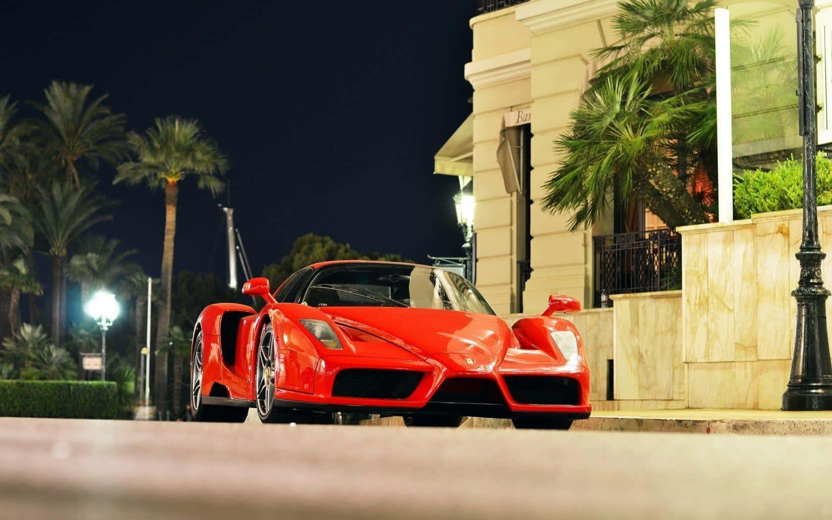 Ferrari Enzo 桌布 壁紙 — 一輛鮮紅色的法拉利Enzo在夜晚停泊在豪華建築前，周圍有棕櫚樹和街燈。