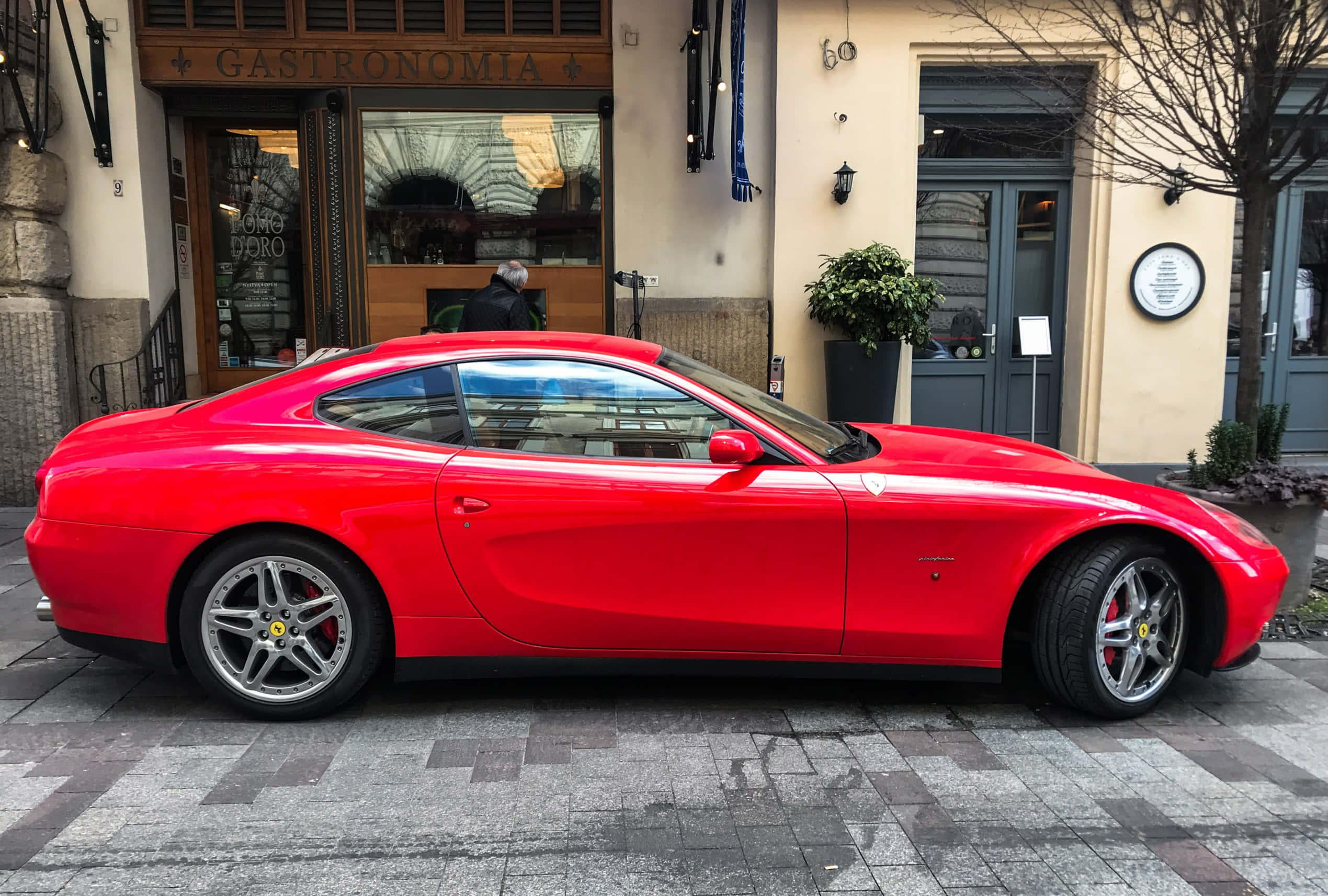 Ferrari 612 Scaglietti 桌布 壁紙 — 一輛鮮紅色的法拉利跑車停泊在濕漉漉的鵝卵石街道上，前方是餐廳，周圍有光禿的樹木和柔和的白日光線。