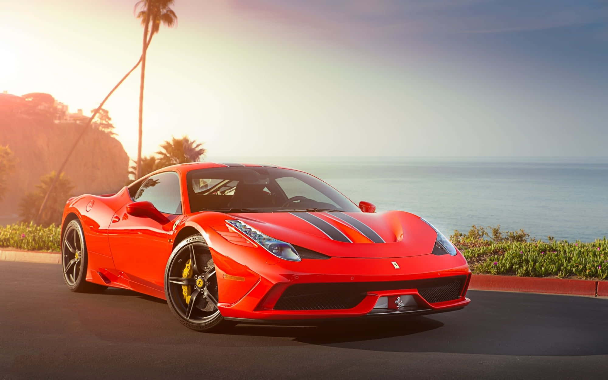 Ferrari 458 Speciale 桌布 壁紙 — 一輛鮮紅色的法拉利跑車在日落時分的海岸公路上閃閃發光，背景有棕櫚樹和海洋。