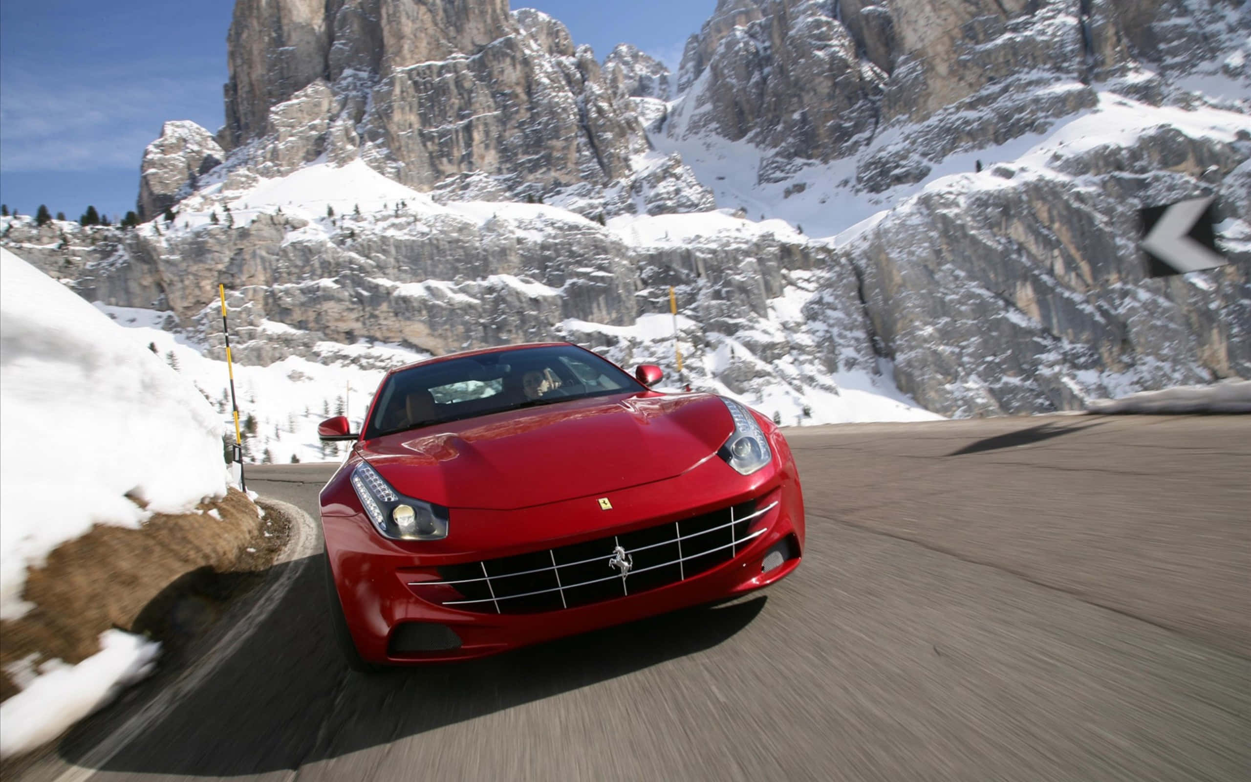 Ferrari Ff 桌布 壁紙 — 一輛鮮紅色的法拉利跑車在晴朗的藍天下，沿著蜿蜒的山路疾馳，兩旁是白雪覆蓋的懸崖。