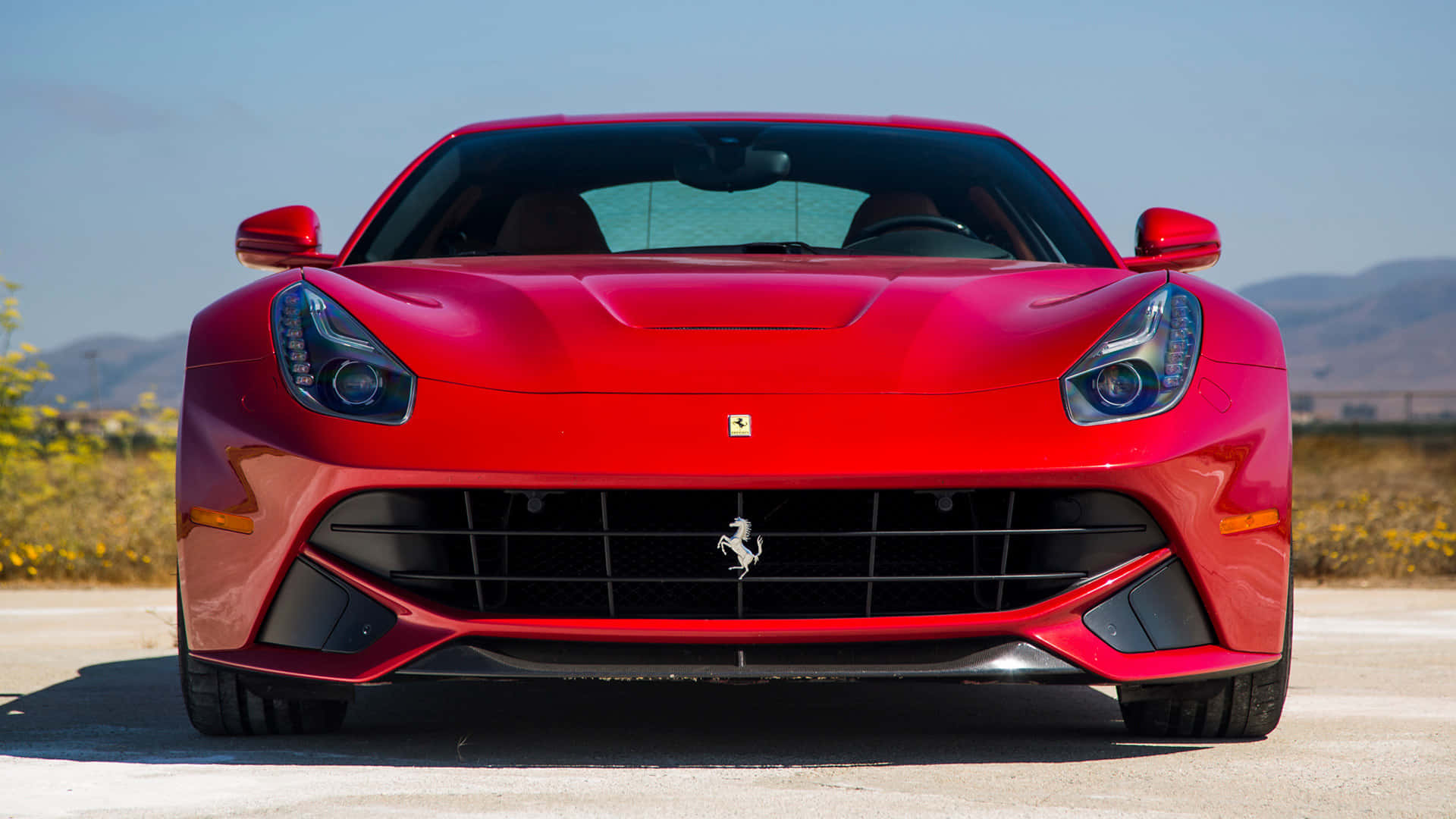 Ferrari F12 Berlinetta 桌布 壁紙 — 一輛鮮紅色的法拉利跑車在陽光明媚的道路上向前行駛，遠處有山丘，天空清澈湛藍。