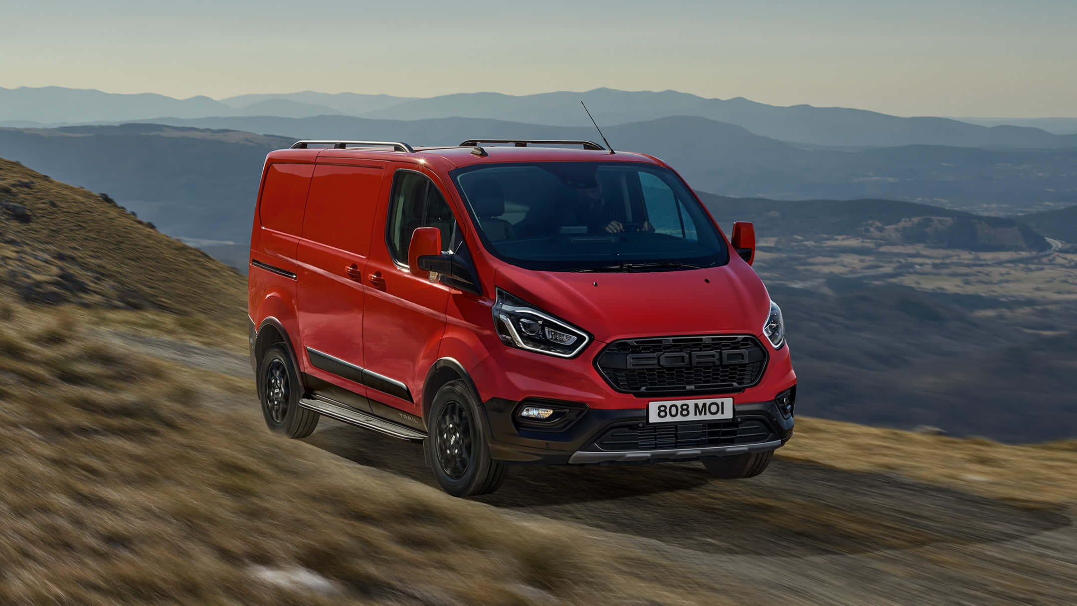 Ford Transit 桌布 壁紙 — 一輛鮮紅色的福特全順客貨車在崎嶇的山路上行駛，背景是模糊的地形和遠處的山峰，天空朦朧。
