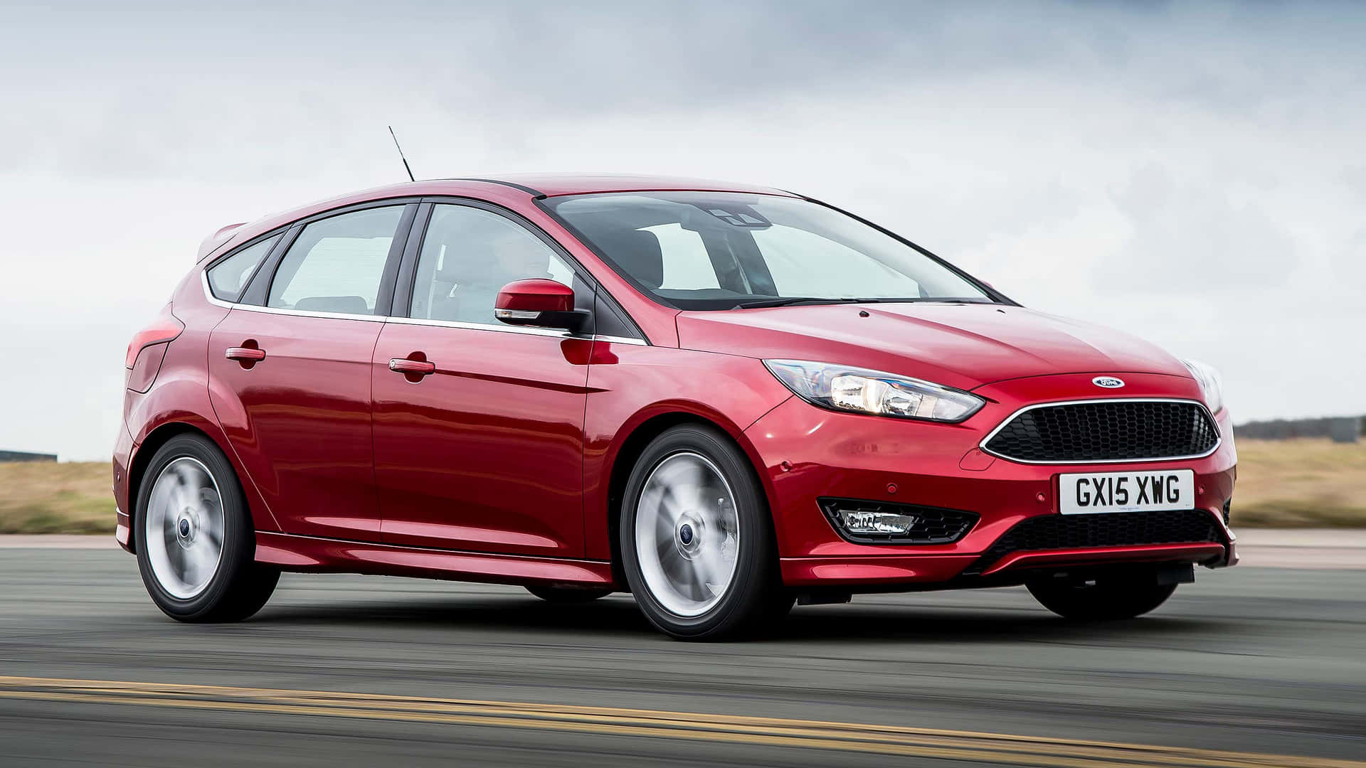 Ford Focus 桌布 壁紙 — 一輛鮮紅色的福特福克斯掀背車在多雲的天空下，沿著模糊的道路疾馳。