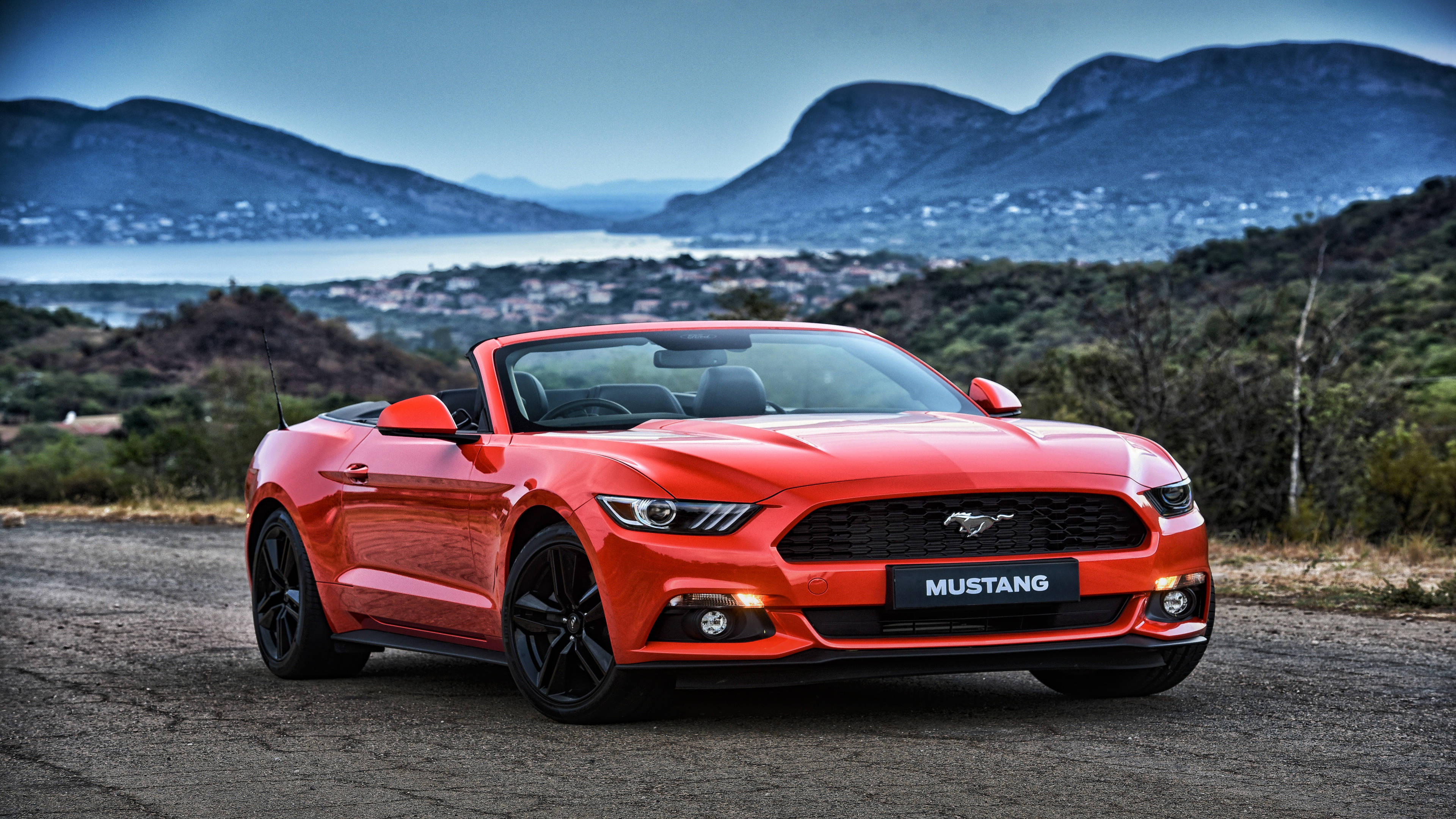 mustang HD 桌布 壁紙 — 一輛鮮紅色的福特野馬敞篷車停泊在碎石路上，背景有山脈和沿海小鎮。