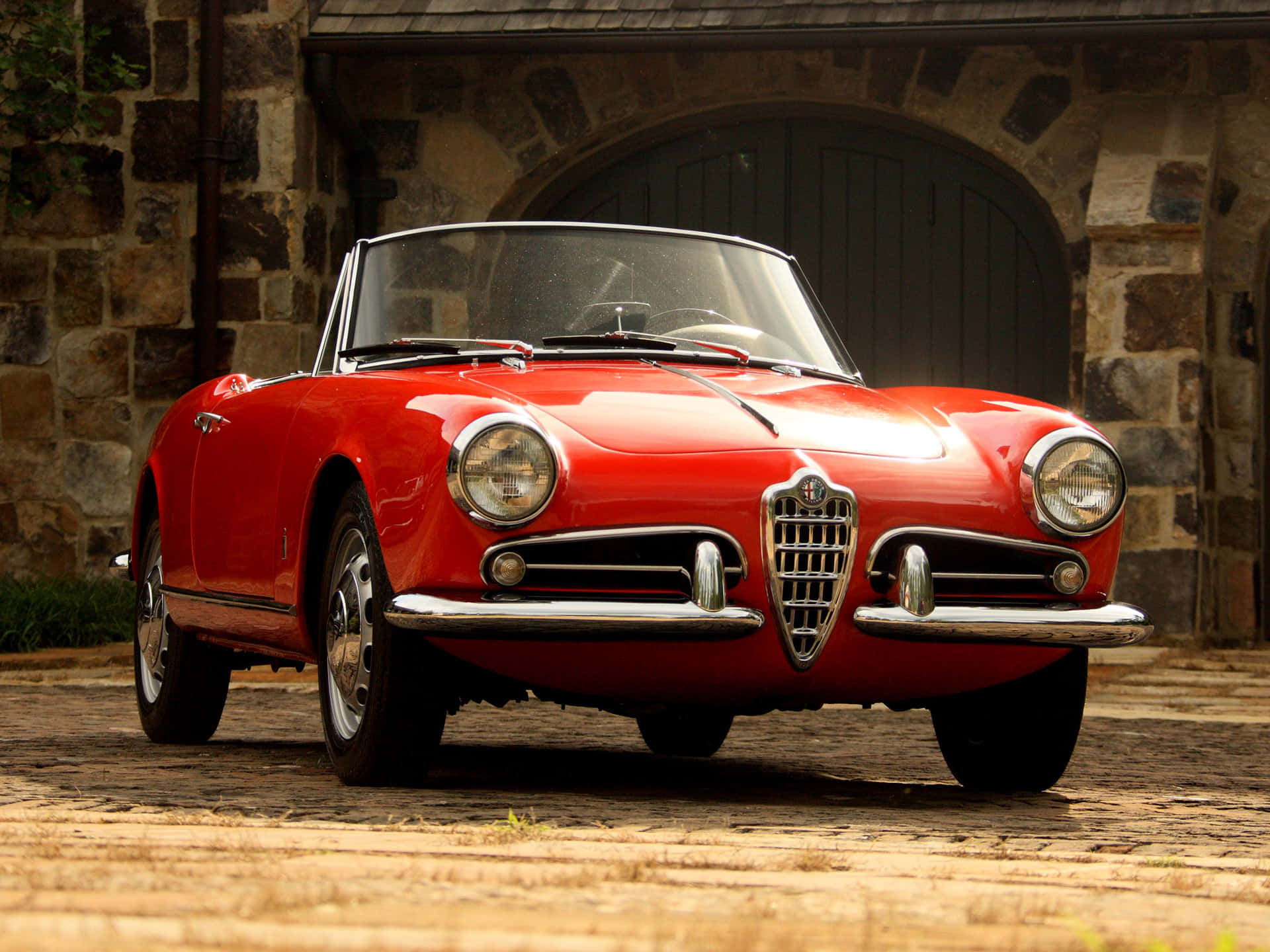 Alfa Romeo Spider 桌布 壁紙 — 一輛鮮紅色的經典愛快羅密歐Spider開篷車停在石砌車庫前的鵝卵石上，車庫有拱形木門。