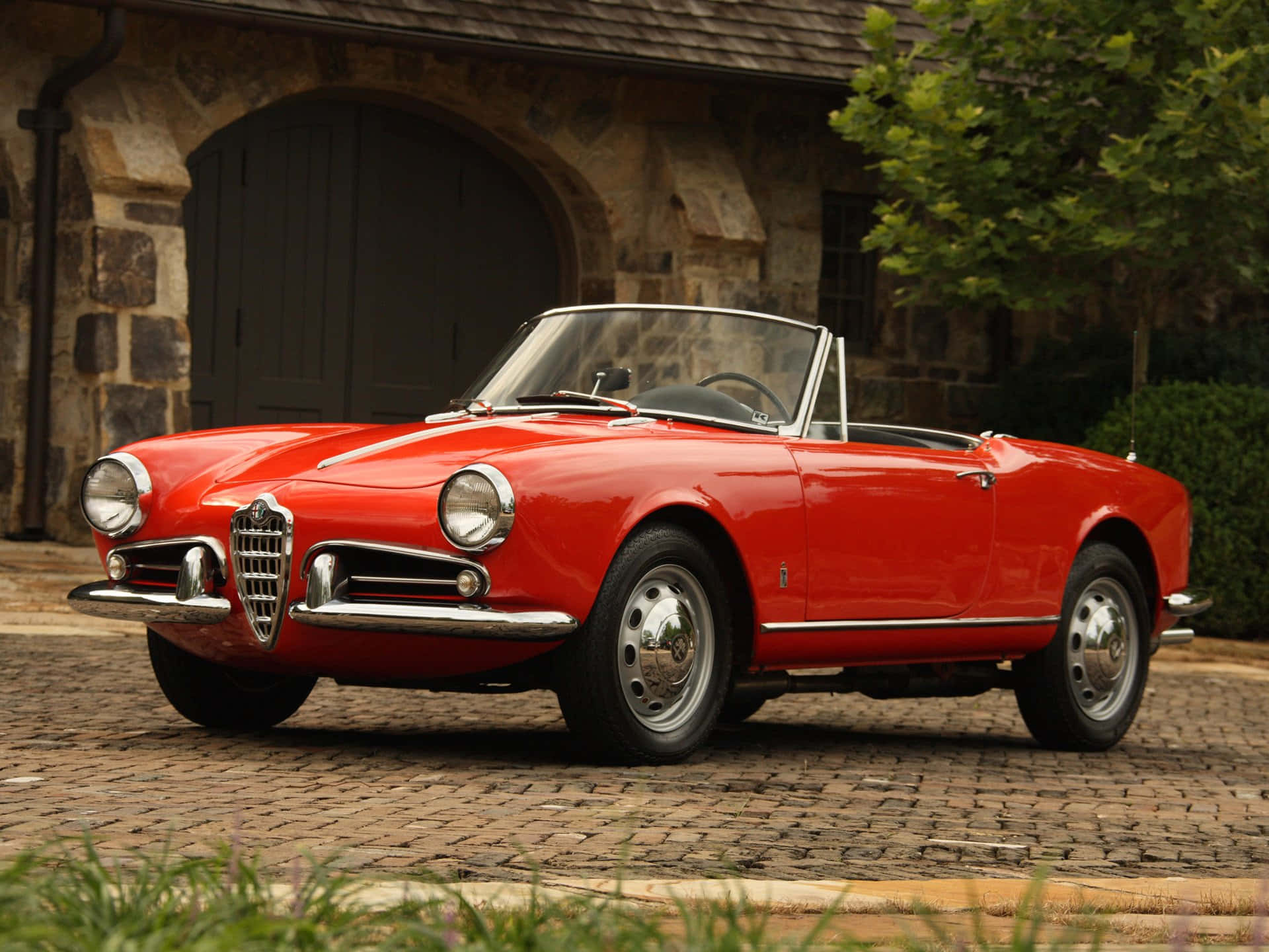 Alfa Romeo Spider 桌布 壁紙 — 一輛鮮紅色的經典愛快羅密歐Spider開篷車停泊在石砌建築前的鵝卵石車道上，周圍有綠植。