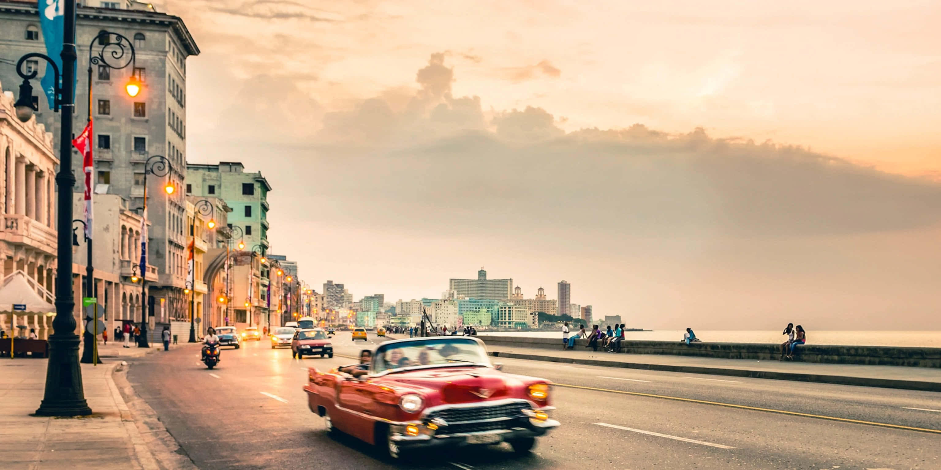Cuba 桌布 壁紙 — 一輛鮮紅色的經典汽車在日落時分沿著夏灣拿的海濱大道行駛，兩旁是歷史建築和閃爍的路燈。