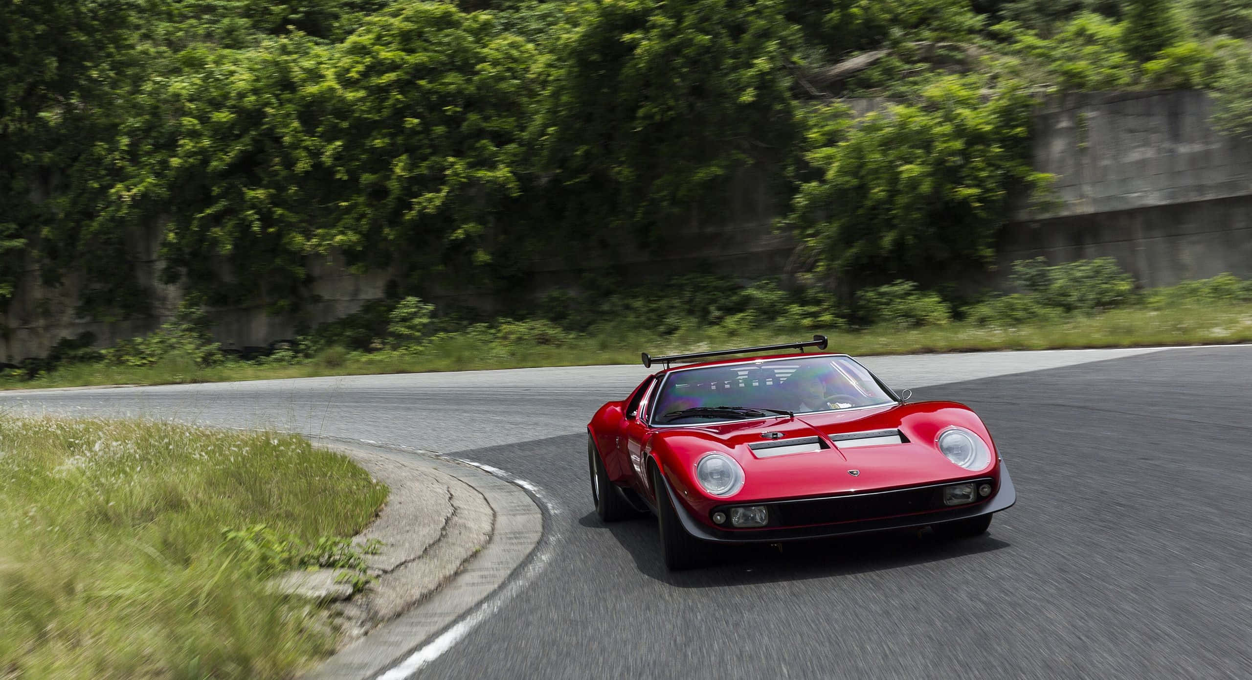 Lamborghini Miura 桌布 壁紙 — 一輛鮮紅色的經典賽車在蜿蜒的山路上疾馳，兩旁綠樹成蔭。