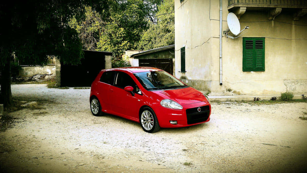 Fiat Punto 桌布 壁紙 — 一輛鮮紅色的緊湊型汽車停泊在塵土飛揚的碎石車道上，旁邊是一座帶有綠色百葉窗的風化米色建築。