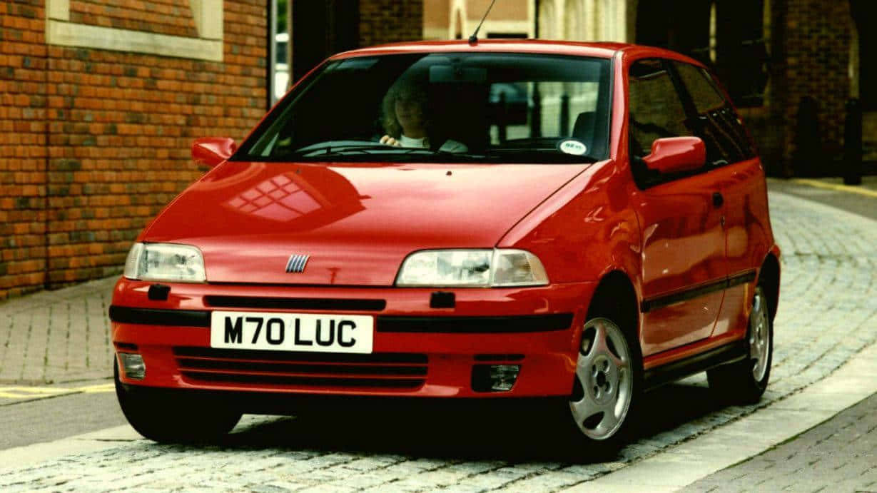 Fiat Punto 桌布 壁紙 — 一輛鮮紅色的菲亞特 Punto 在日光下停泊在磚砌建築旁的鵝卵石街道上。