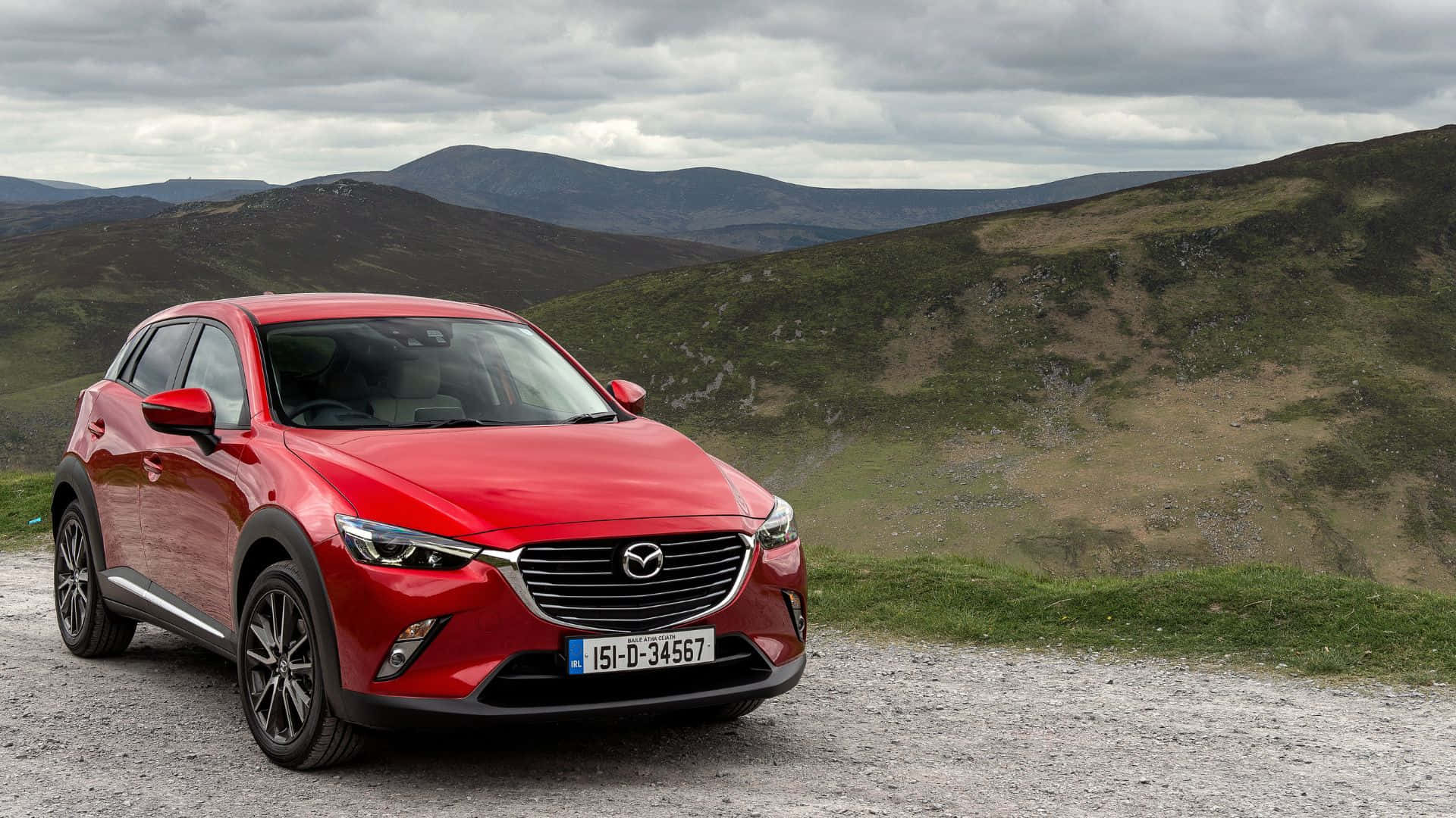 Mazda Cx-3 桌布 壁紙 — 一輛鮮紅色的萬事得CX-3 SUV停泊在碎石路上，背景是起伏的綠色山丘和陰天。