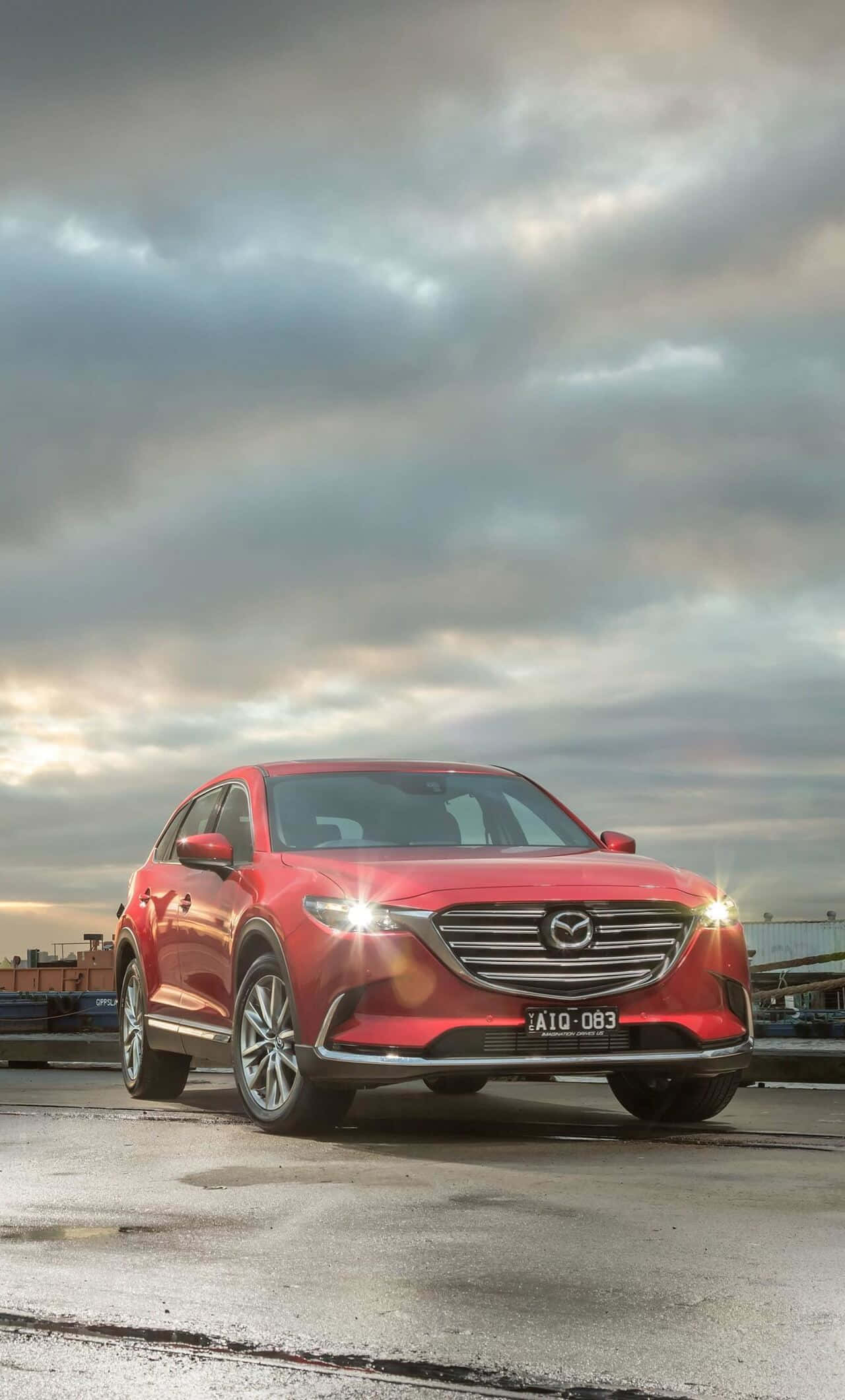 Mazda Cx-9 桌布 壁紙 — 一輛鮮紅色的萬事達CX-9 SUV在戲劇性的多雲天空下閃閃發光，車頭燈亮著，停在濕漉漉的工業碼頭上。