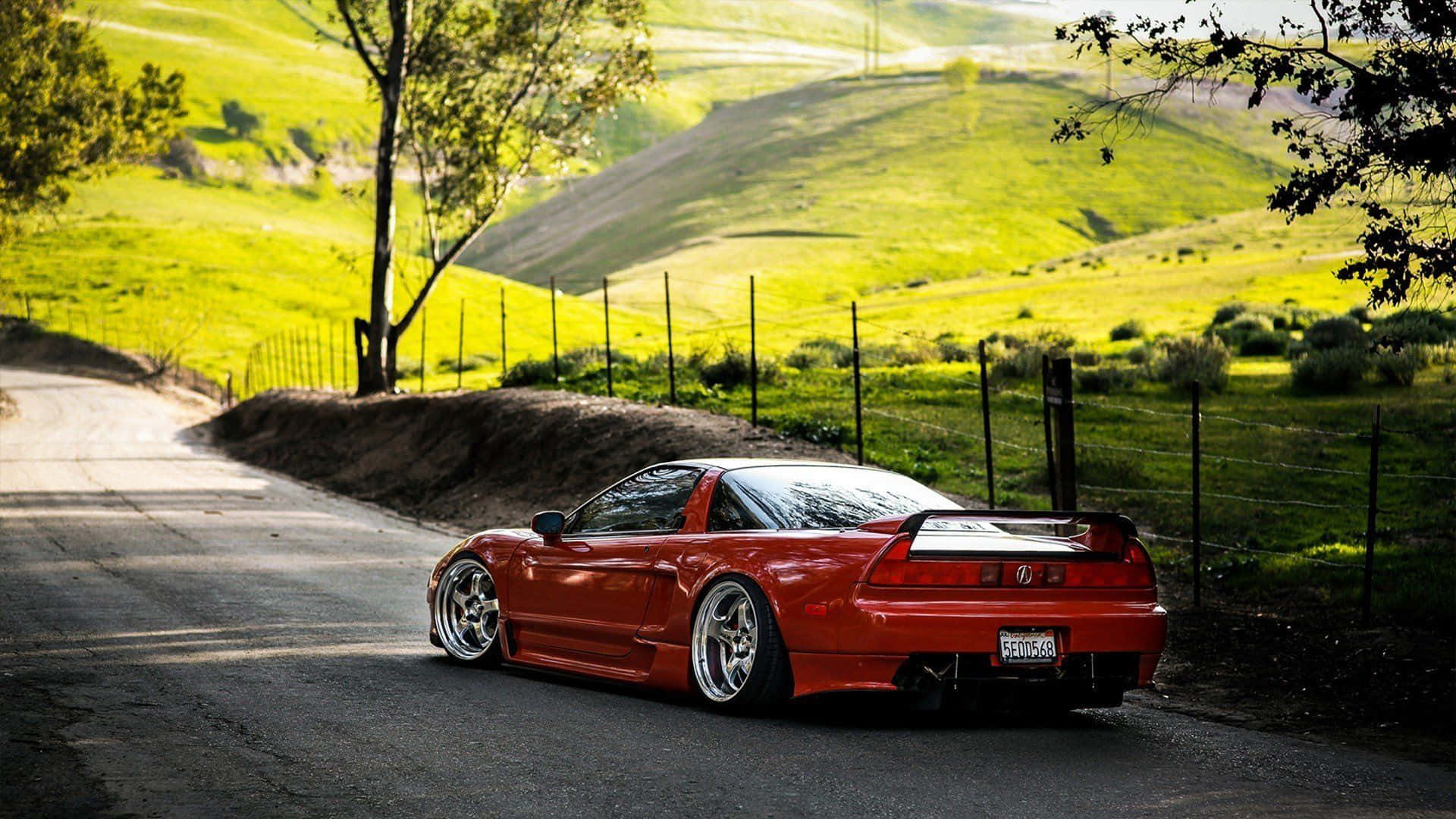 JDM 車 桌布 壁紙 — 一輛鮮紅色的跑車停泊在蜿蜒的鄉村道路上，周圍是陽光照耀的綠色山丘和樹木。