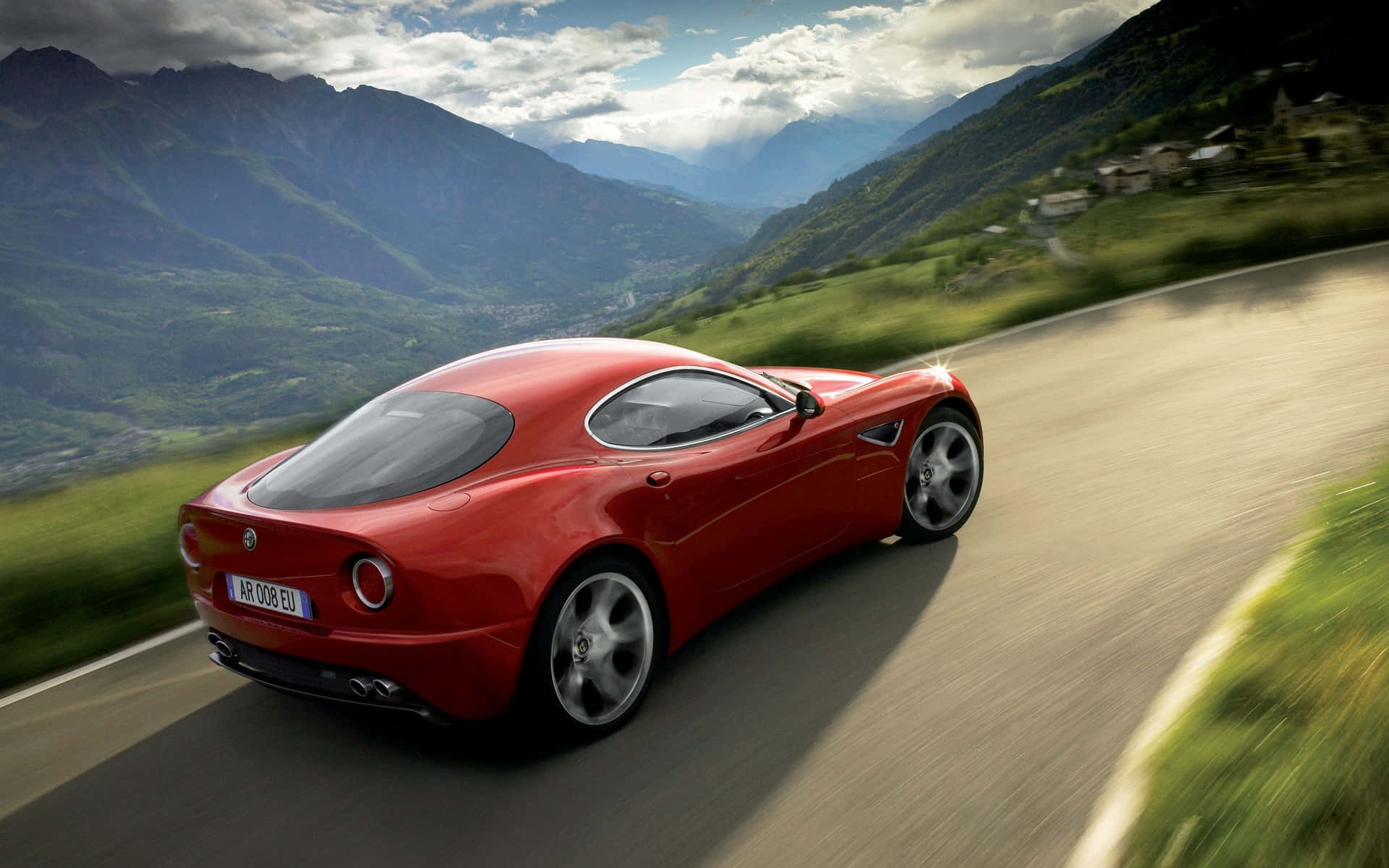 Alfa Romeo 8c Competizione 桌布 壁紙 — 一輛鮮紅色的跑車在蜿蜒的山路上疾馳，兩旁是翠綠的山谷和遠處的村莊，天空部分多雲。