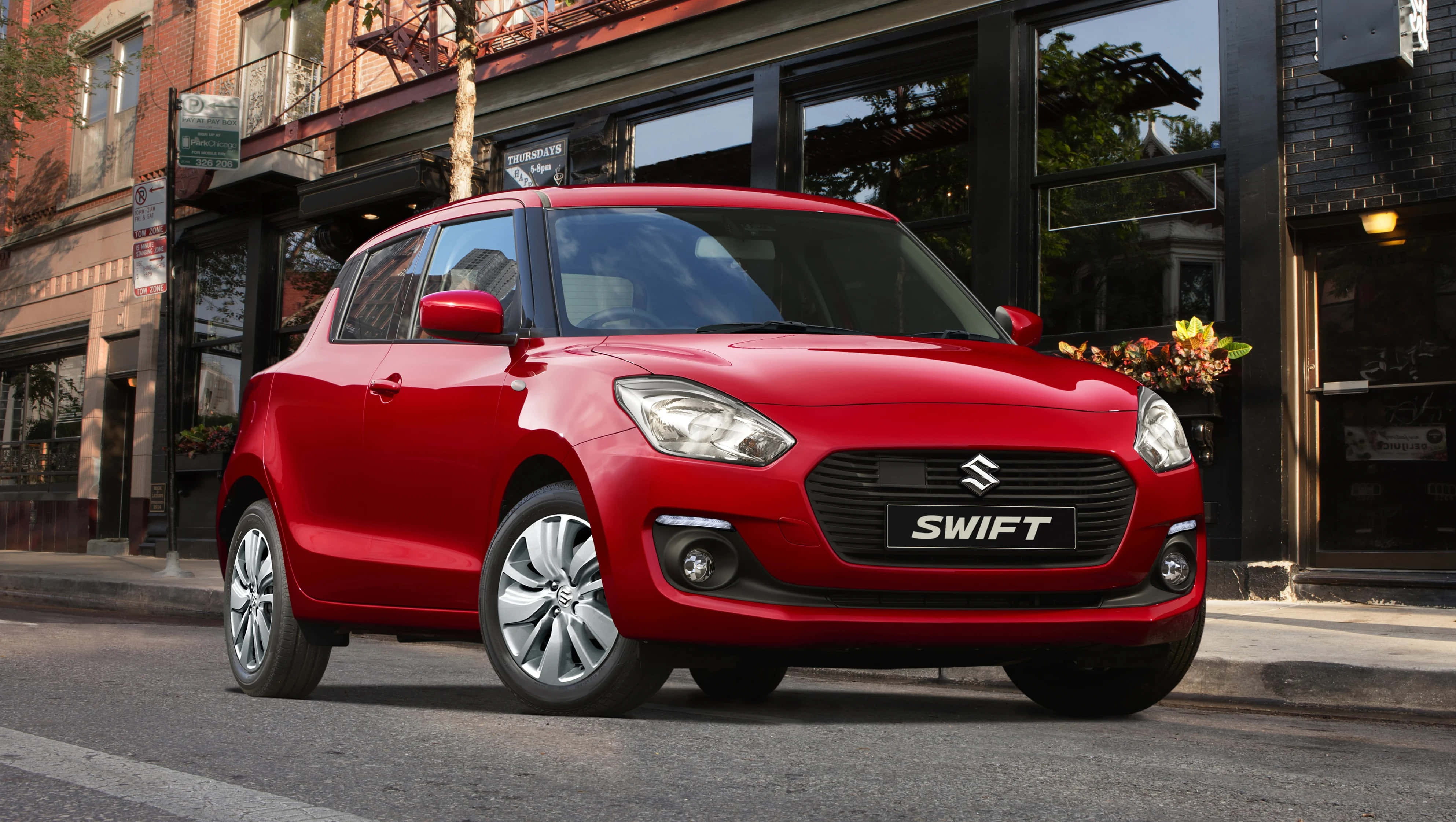 Compact Car 桌布 壁紙 — 一輛鮮紅色的鈴木 Swift 停泊在城市的街道上，背景是磚砌建築和商店門面。