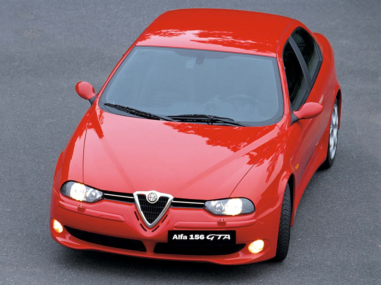Alfa Romeo 156 桌布 壁紙 — 一輛鮮紅色的阿爾法156 GTA跑車在日光下閃耀，車頭燈亮著，停在灰色瀝青路面上。