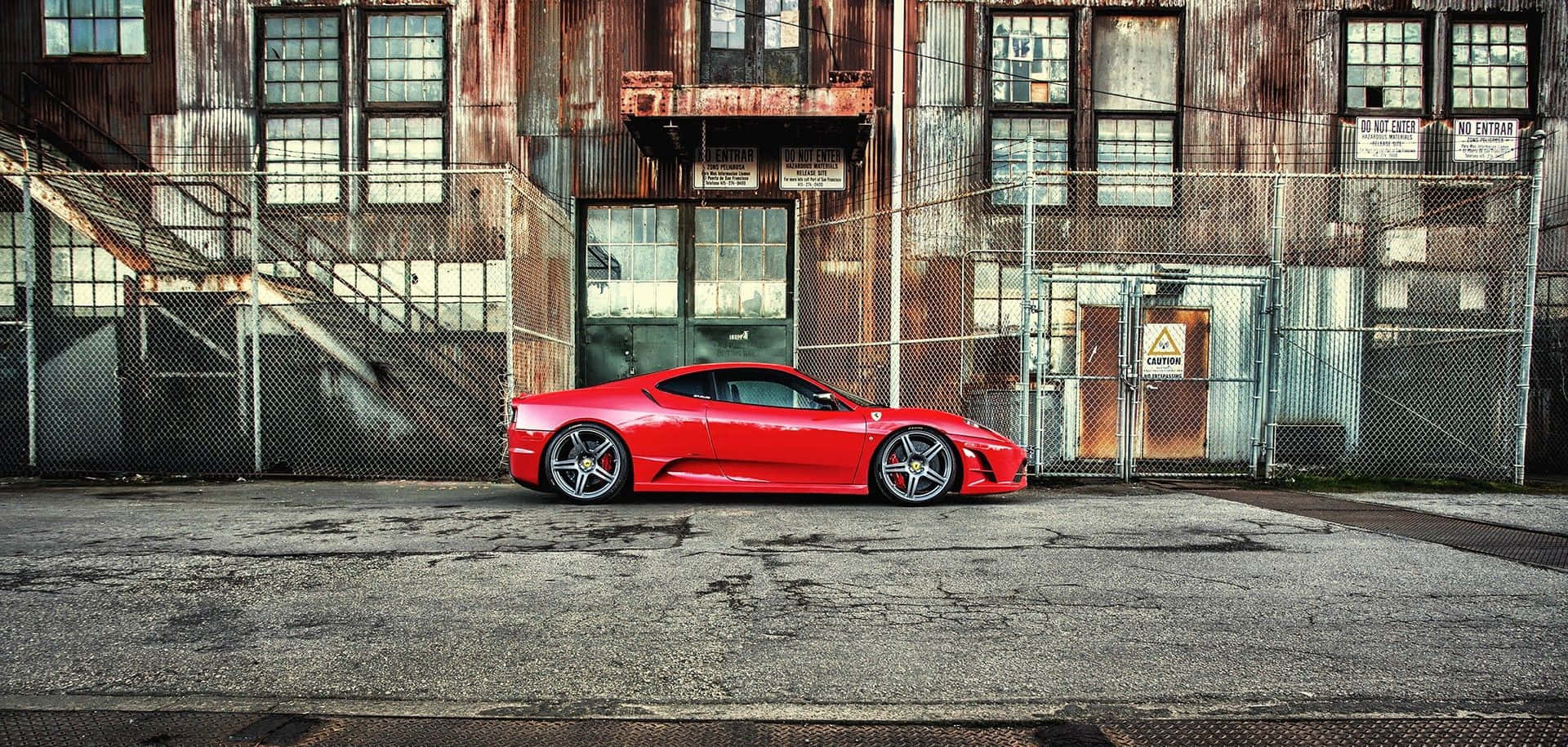 Ferrari 360 Modena 桌布 壁紙 — 一輛鮮紅色跑車停泊在風化嘅工業建築前面，周圍有生鏽金屬同鐵絲網圍欄。