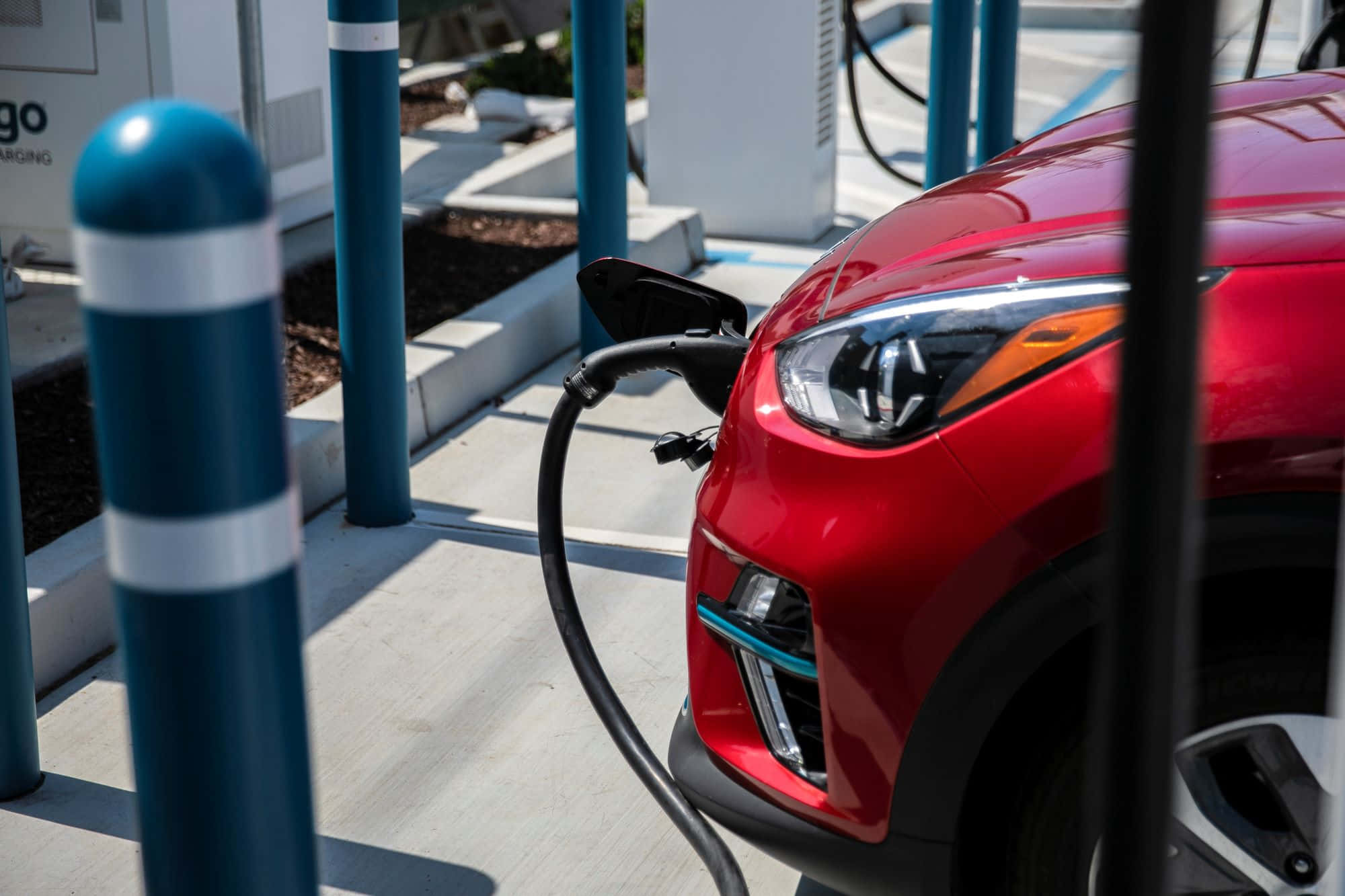 Electric Vehicle 桌布 壁紙 — 一輛鮮紅色電動車在明亮的日光下連接到充電站，前景有藍色護柱。